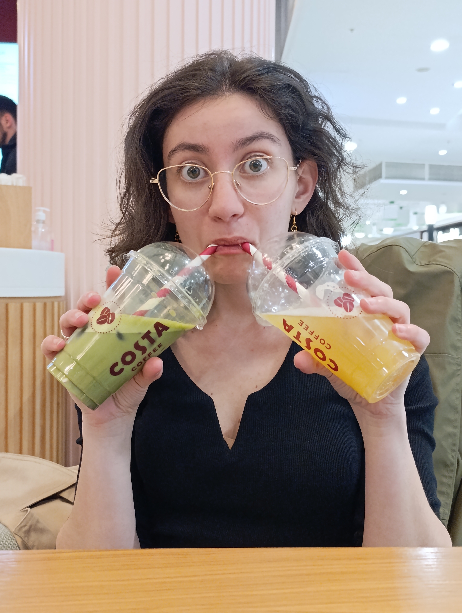 A young woman is captured in a close-up, eye-level shot, playfully attempting to drink from two Costa Coffee cold beverage cups at once. She has dark, curly hair and wears distinctive gold-rimmed round glasses and gold drop earrings. Her wide-open eyes and slightly puckered lips, with a red-striped straw from each cup inserted, convey a humorous and surprised expression as she looks directly at the camera.
In her left hand, she holds a transparent cup filled with a bright green beverage, likely matcha, with the "COSTA COFFEE" logo printed vertically in red letters. Her right hand grasps a similar transparent cup containing a vibrant yellow or orange drink, possibly a fruit juice, also featuring the "COSTA COFFEE" branding. Both cups are topped with clear domed lids and red-striped straws.
The woman is wearing a black, v-neck ribbed top. A warm-toned wooden table occupies the immediate foreground. The background features light-colored, possibly pink, vertical paneling or pillars, suggesting an indoor public space like a café or food court. A blurred figure of a man is partially visible in the far left background, and a beige jacket is draped over a chair or her shoulder on the right. The bright artificial lighting indicates an indoor setting, possibly in Sofia, Bulgaria, as per the provided context.