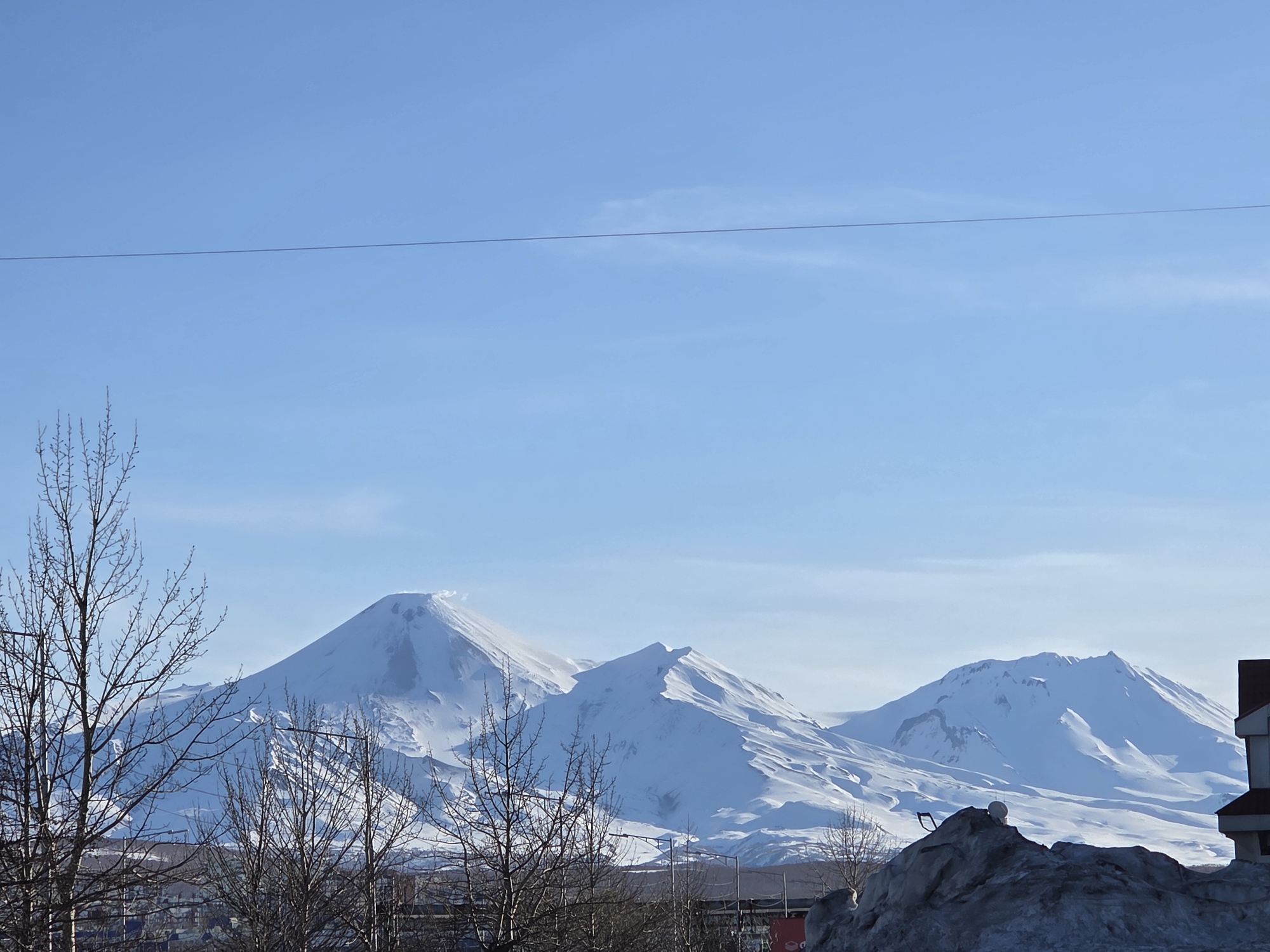 The image depicts a daytime scene with three snow-covered mountains against a clear, light blue sky. In the foreground, there are bare trees and a pile of snow. In the background, a building is partially visible. The mountains are the most prominent feature, with the central peak appearing somewhat conical and potentially volcanic in nature. The scene appears to be located near the city, since part of a residential building is on the right side. The location is Unknown, Unknown.