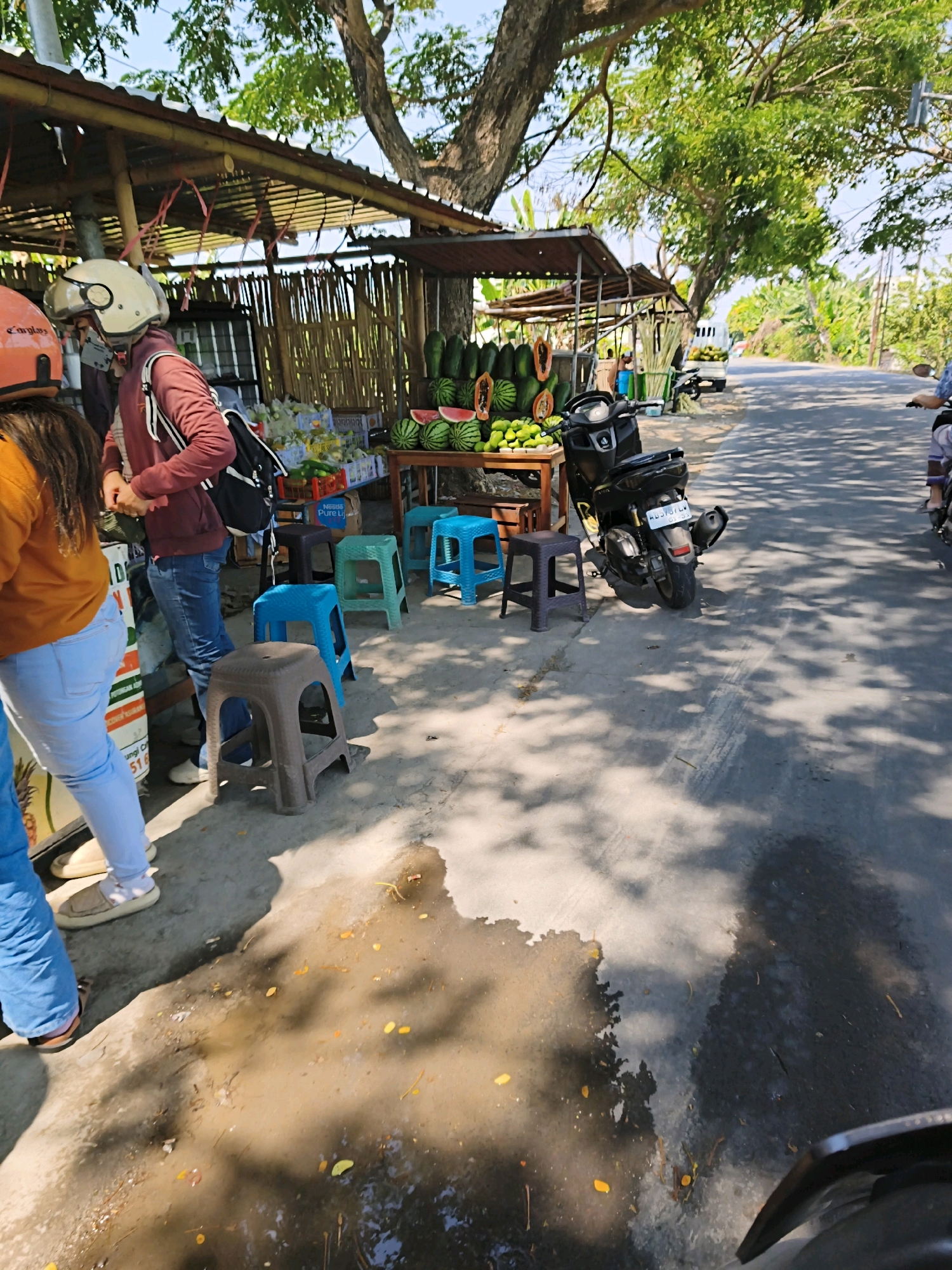 This image depicts a roadside fruit stand in Desa Pandeyan, Indonesia, on a sunny day. A covered stall with a bamboo-like structure and a corrugated metal roof displays various fruits including watermelons, papayas, and what appear to be mangoes or similar tropical fruits. Several plastic stools of different colors (blue, green, and brown) are arranged in front of the stall. A black motorcycle is parked near the stall. On the left side of the image, a person wearing a white helmet and a maroon jacket is standing, and another person in an orange top and blue jeans is partially visible. A road with passing traffic is visible in the background. The ground in front of the stall is a mixture of concrete and dirt, with puddles of water and fallen leaves. The overall scene suggests a typical rural or semi-rural roadside market in Indonesia.