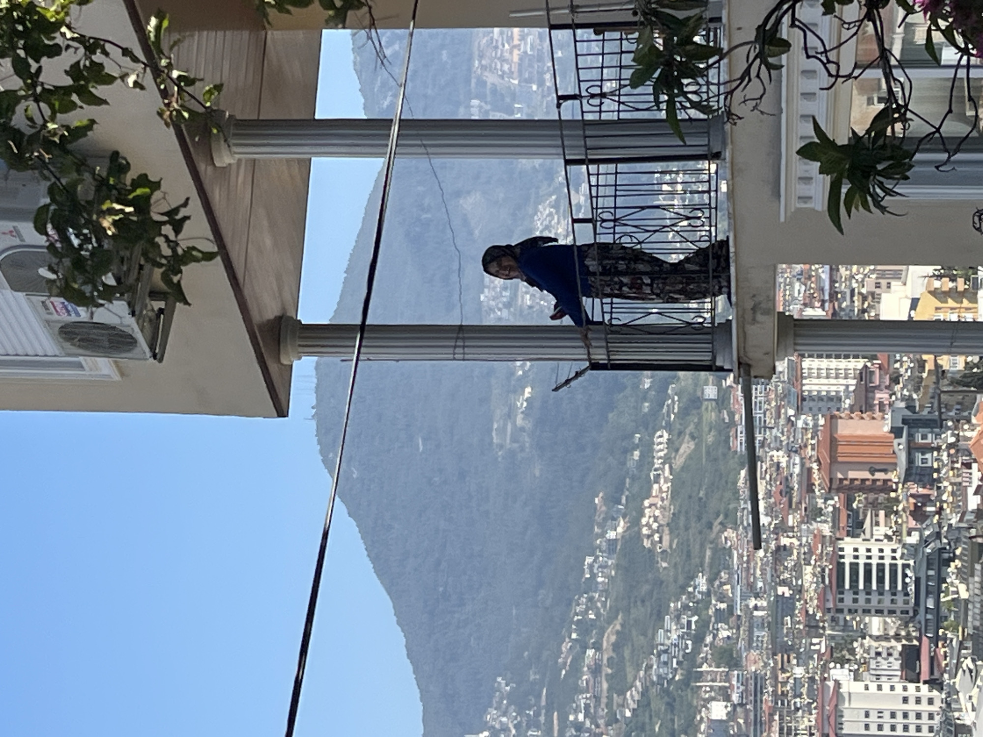 A daytime photograph, originally rotated 90 degrees clockwise, captures a woman standing on a balcony with a panoramic backdrop of a city nestled against mountains. The woman, smiling and looking towards the camera, wears a blue top, patterned trousers, and a head covering. She is holding a long pole or stick.
The balcony features white, fluted columns and a black, decorative metal railing. To the left (after rotation), an outdoor air conditioning unit, with "Mitsubishi" visible on its casing, is mounted on the building wall. Green foliage from plants hangs into the frame from the top left and right corners.
Beyond the balcony, a sprawling city extends across a valley or hillside, filled with numerous buildings. In the mid-ground, a large, verdant mountain rises prominently, showcasing a rugged natural landscape. The sky above is clear blue, indicating bright, sunny weather. The scene suggests a high vantage point overlooking a city with significant natural surroundings, consistent with the coastal mountainous region of Alanya, Turkey.