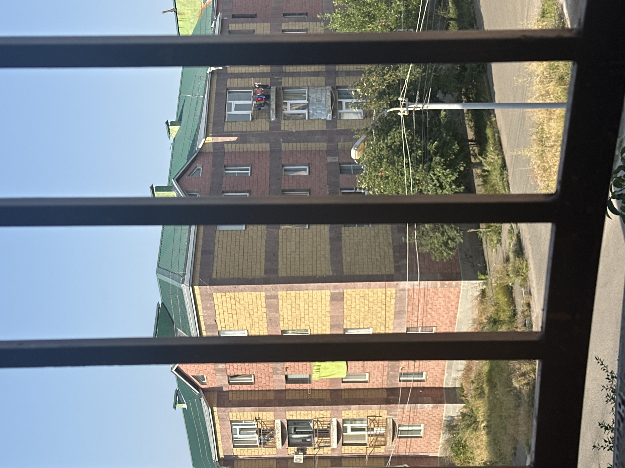 This is an exterior, eye-level shot of a multi-story apartment building in Gyumri, Armenia, viewed through a dark metal grate. The building features a predominantly yellow brick facade with red brick accents, and a green tiled roof. Several balconies are visible, some with laundry drying on them. The ground in front of the building is covered with dry grass and weeds. A street and a utility pole with overhead wires are also present. The sky is clear and blue.
