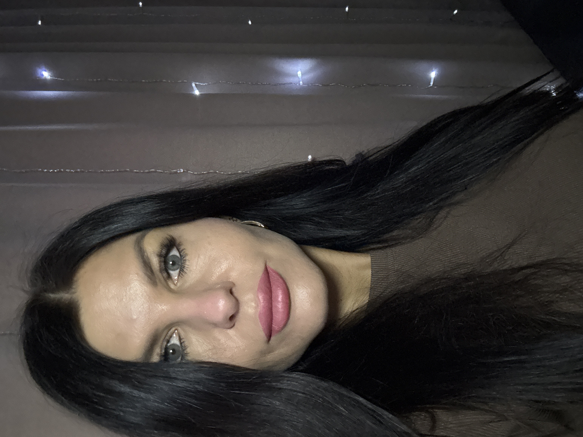 The image is a close-up portrait of a woman with long, black hair. She has fair skin, gray eyes, and pink lipstick. She is wearing a brown top. In the background, there are out-of-focus string lights, suggesting an indoor setting. The image does not provide enough information to determine if it was taken in the city of Chaikovsky, Russia.