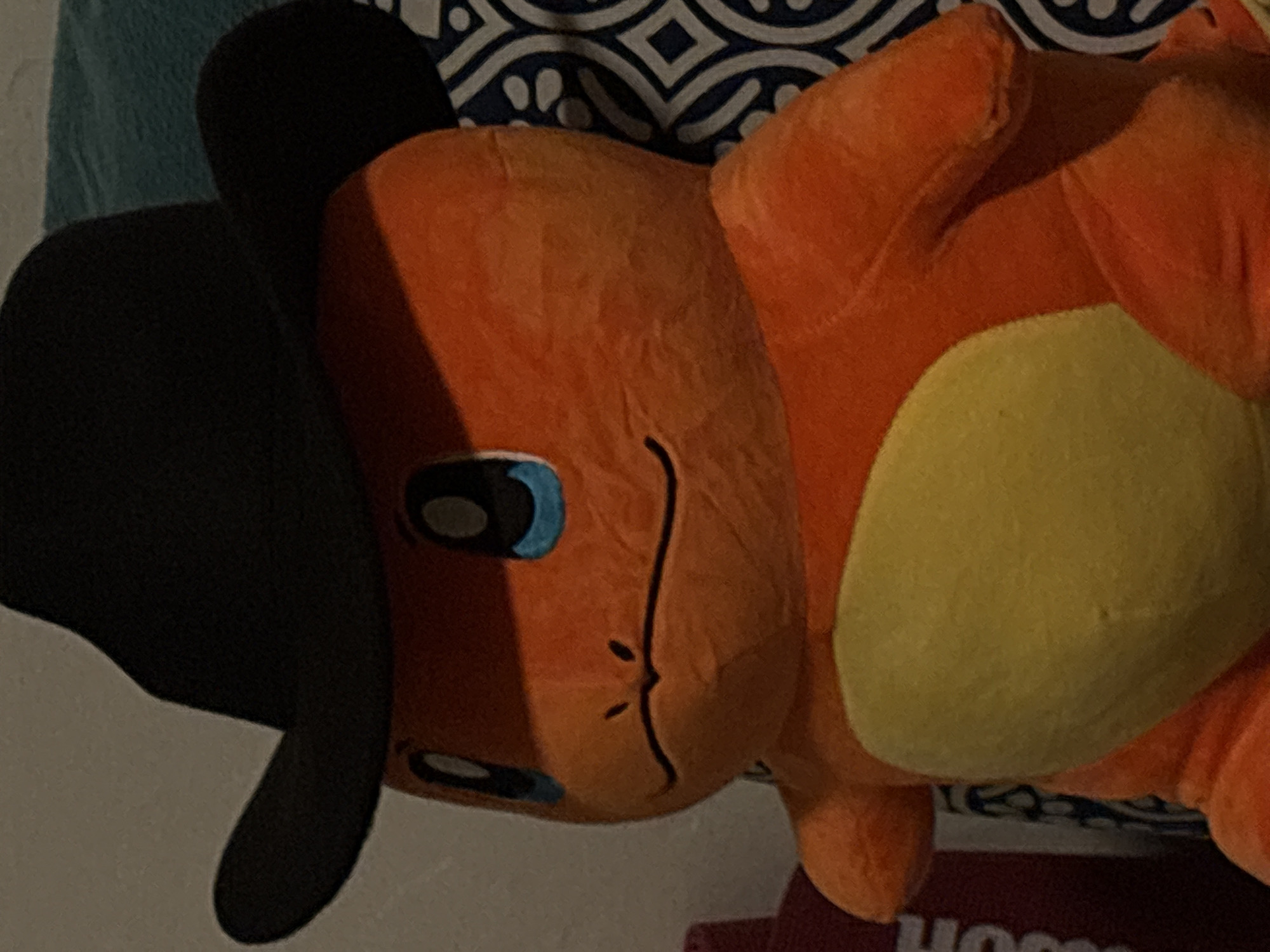 The image shows a plush toy resembling the Pokémon character Charmander wearing a black hat. The toy has orange fur, a yellow stomach, and blue and black eyes. It is lying on what appears to be bedding, which includes a teal pillow and a patterned pillow with dark blue and white designs. The lighting is uneven, casting shadows across the toy's face. The location is not discernible from the image.