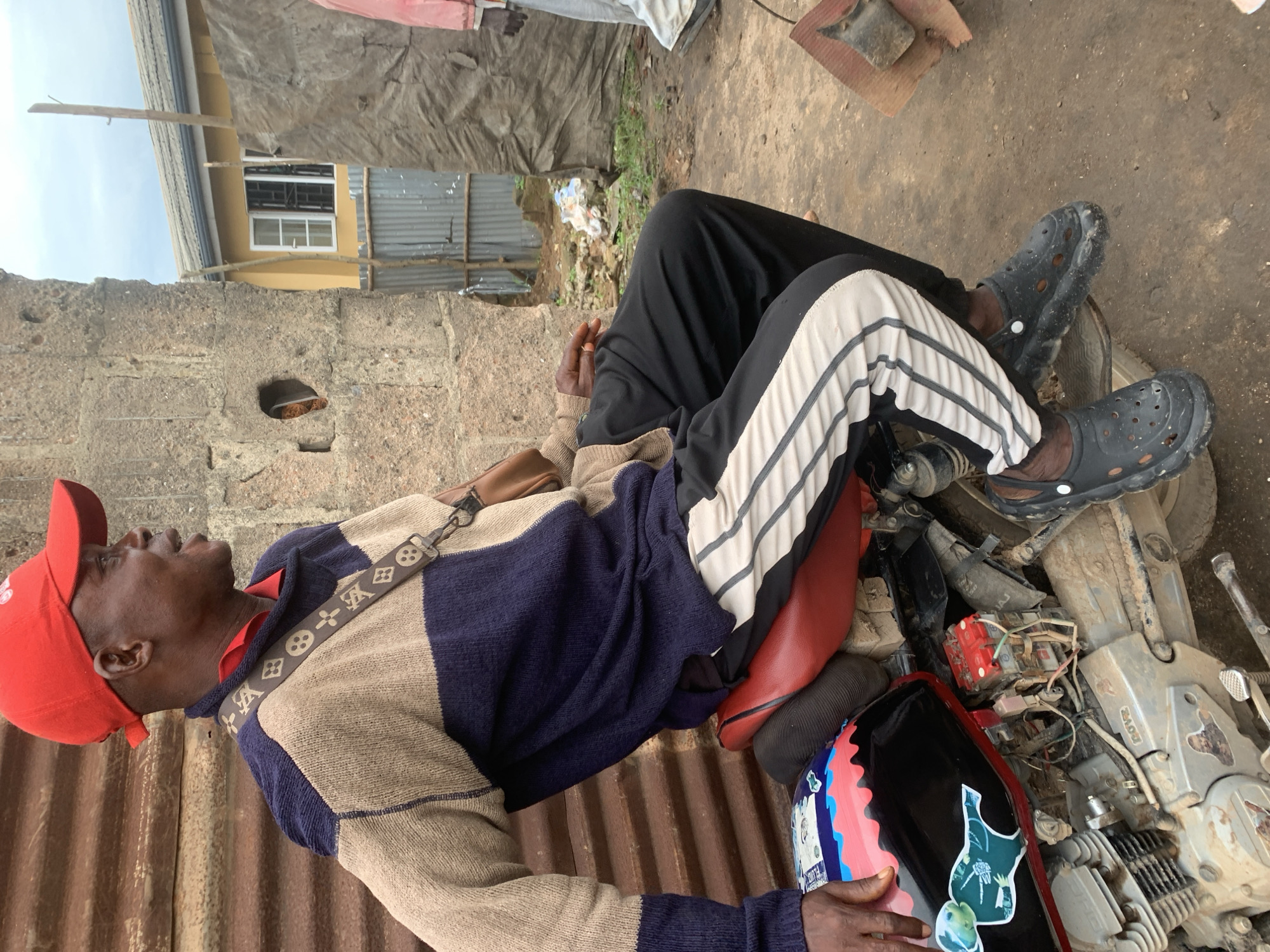 A Black man wearing a red cap, a blue and tan sweater, and black pants with white stripes is sitting on a red motorcycle. He has a brown crossbody bag with a patterned strap. The motorcycle has a black and red fuel tank with colorful graphics. The background shows a textured wall, corrugated metal sheeting, a yellow building with a window, and a tarp. The ground is dirt. The image appears to be taken in Shagamu, Nigeria, a city known for its pottery and brick manufacturing, which might explain the rustic, unpaved environment and the types of structures visible.