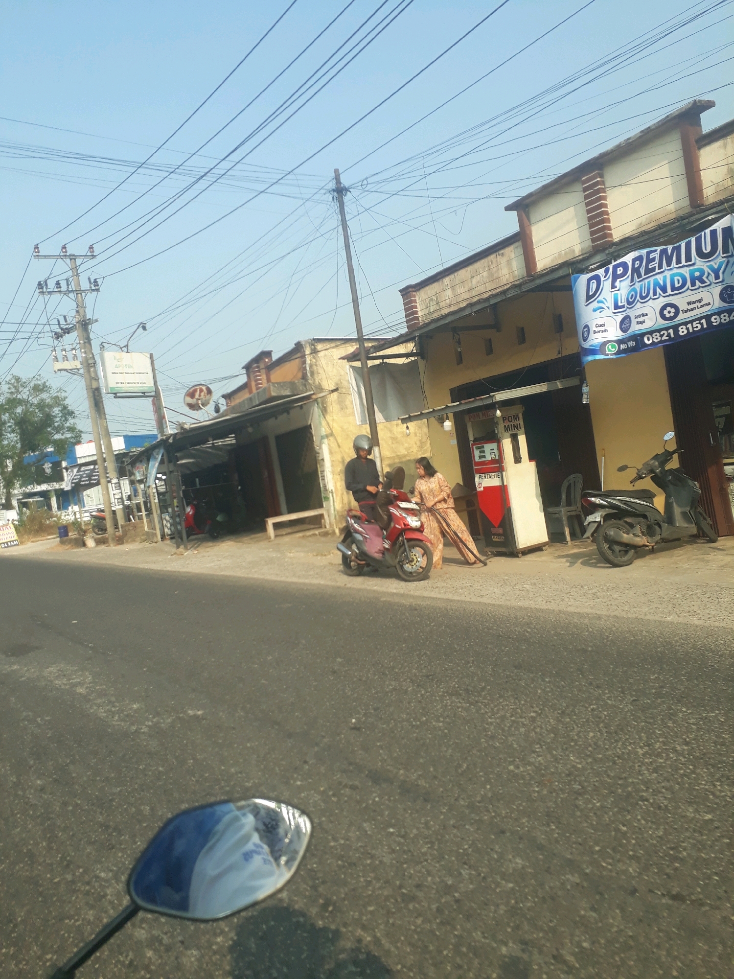 The image captures a street scene in Pangkalpinang, Indonesia, with several shops lining the road under a clear blue sky. In the foreground, the asphalt road is visible, with a motorcycle's mirror reflecting the surrounding environment, including a portion of a white garment.
On the right side of the street, a building with a yellow facade and brown trim features a prominent banner for "D'PREMIUM LAUNDRY," advertising services like "Cuci Bersih," "Setrika Rapi," and "Wangi Tahan Lama" (Clean Wash, Neat Ironing, Long-lasting Fragrance). The banner also includes a phone number: "0821 8151 984" and "No Wa". Adjacent to the laundry is a "POM MINI" (mini gas station) with a red and white pump, labeled "PERTALITE". A woman in a patterned dress is standing next to the gas pump, possibly interacting with it or the attendant. A man wearing a black shirt and helmet is seated on a red scooter, to the left of the gas station. Another black scooter is parked to the right of the laundry building.
Further down the street, there are more shopfronts, one of which has a sign reading "APOTIK" (pharmacy), with text indicating it sells "OBAT-OBAT DAN ALAT KESEHATAN" (medicines and health equipment). Electrical poles with numerous wires crisscross above the scene, typical of urban infrastructure. Trees and foliage are visible on the left side of the street. The overall impression is a busy commercial area on a sunny day.