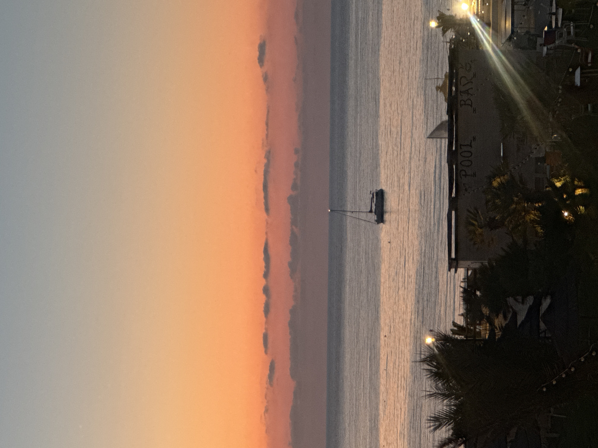 The image depicts a serene sunset over the sea in Isola delle Femmine, Italy. The sky transitions from a pale blue at the top to a vibrant orange and pink near the horizon. A few scattered clouds are visible in the middle distance. A sailboat is anchored on the calm water, appearing as a dark silhouette against the luminous sky. In the foreground, the silhouette of palm trees and structures of a beachfront establishment are visible. One structure has the words "POOL BAR" painted on its wall. Lights are visible on the establishment, suggesting it is either evening or preparing for nighttime operations. The overall atmosphere is peaceful and tranquil.