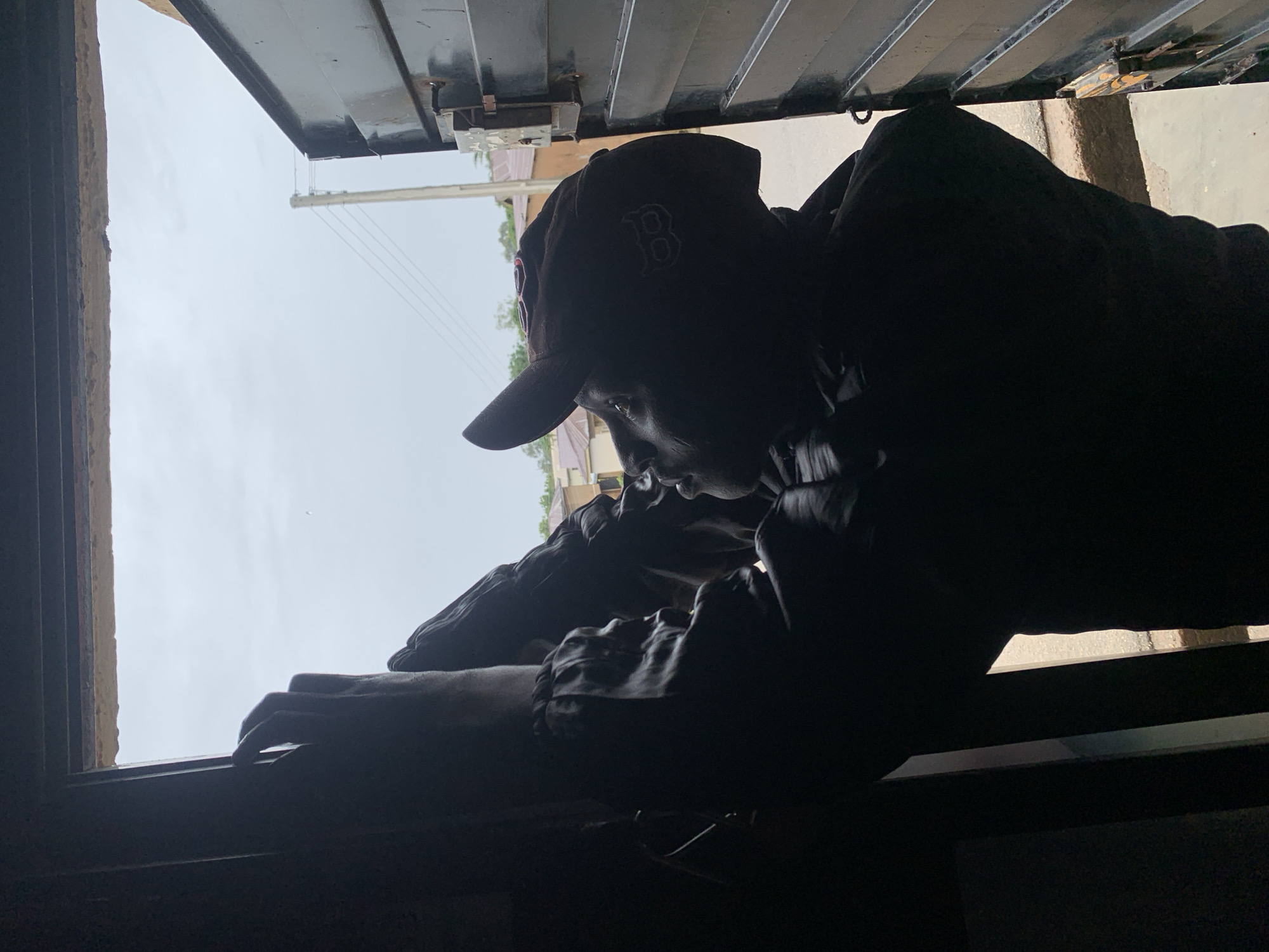 A person wearing a cap and dark clothing is silhouetted against an overcast sky, reaching upwards with their right hand. They appear to be standing within a doorway or opening, looking towards the sky. To the right, a dark metal gate is partially open, revealing a street with buildings and utility poles in the background, suggesting an urban setting. The overall lighting indicates it is daytime, likely in Jos, Nigeria, given the architectural style and environment. The person's action suggests they are attempting to reach or manipulate something outside the opening.