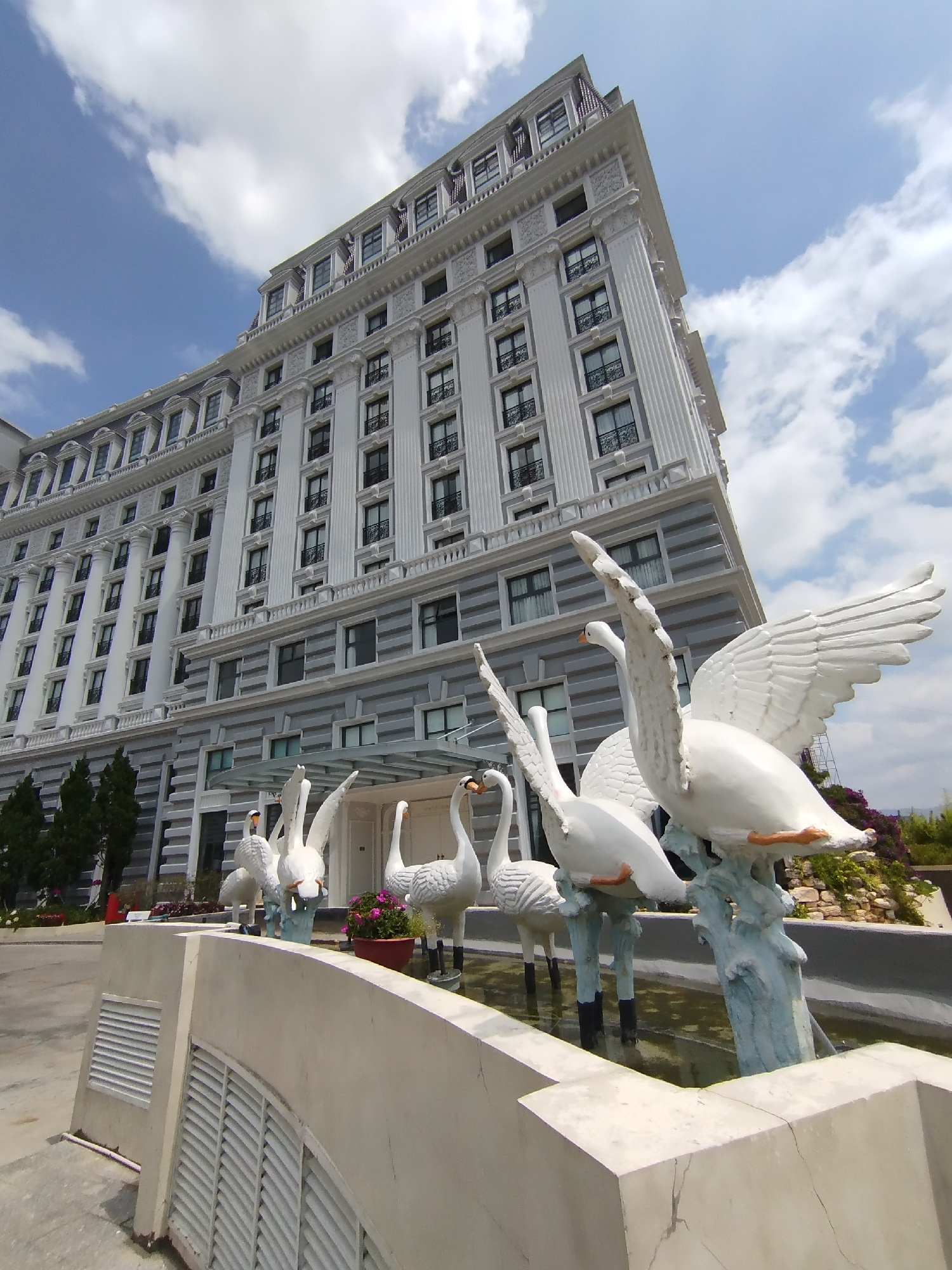 The image shows a large, white, multi-story building in Da Lat, Vietnam. The building features neoclassical architectural elements, including columns, window details, and a cornice. In the foreground, a series of white swan statues stand in a low, water-filled basin. A low wall with ventilation grates borders the basin. The sky is blue with scattered white clouds.