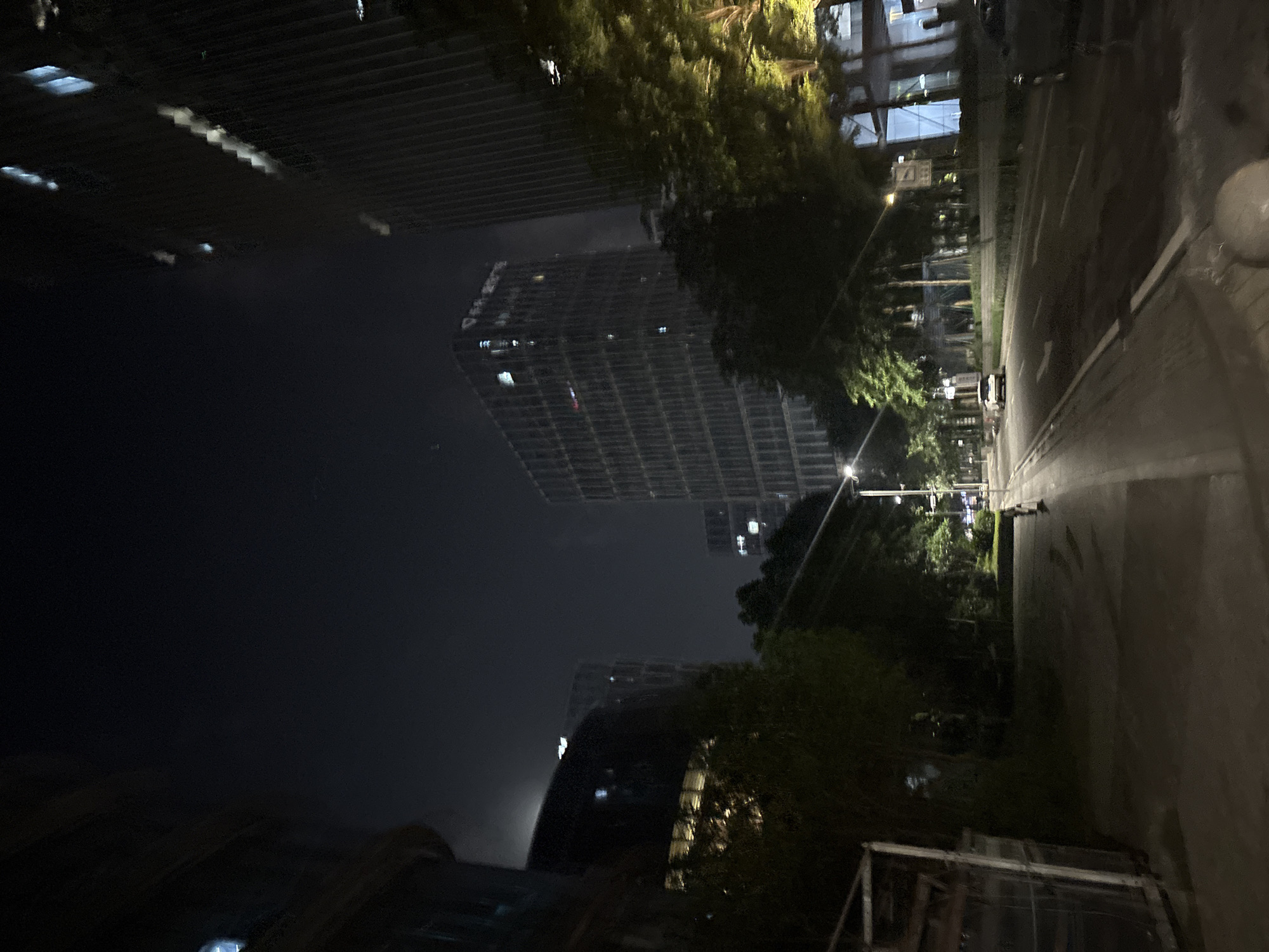 The image depicts a nighttime street scene in the Haizhu District of China, likely a commercial or business area. Tall, modern buildings with illuminated windows dominate the background, their facades reflecting the dim ambient light. Trees line the street, their foliage casting shadows and creating a dappled effect on the asphalt. A car is visible driving down the road, its headlights illuminating the path ahead. The street is wet, suggesting recent rain, and streetlights cast pools of yellow light. The overall impression is one of urban nighttime activity, with a sense of quietness and a slightly melancholic atmosphere due to the darkness and the wet surfaces.