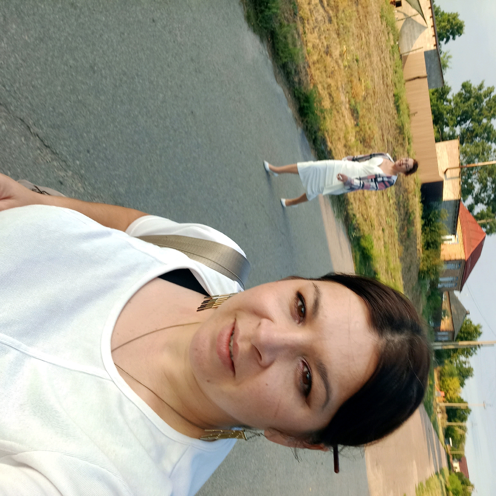 In an outdoor, low-angle selfie, a woman in a white t-shirt is in the foreground, her face turned towards the camera. She is wearing a necklace and large, rectangular earrings. Behind her, on a paved road, another woman in a white dress and a patterned cardigan is walking away from the camera. The background features a grassy incline, trees, and houses, suggesting a rural or suburban setting. The image is oriented sideways.