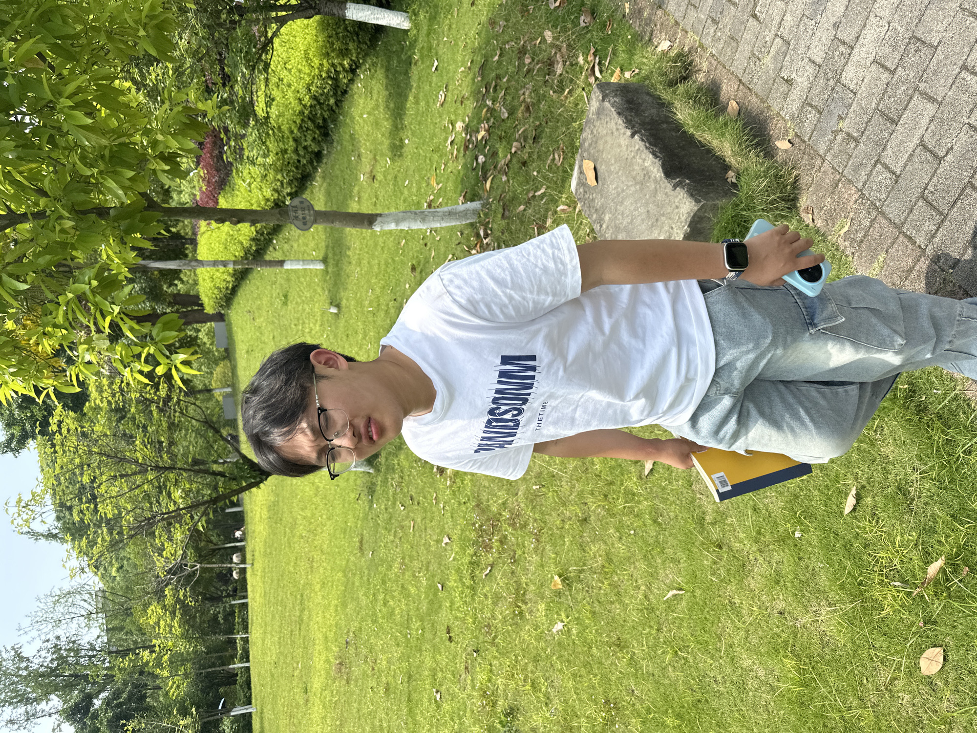 A person stands on a grassy lawn with sparse fallen leaves, a pathway of rectangular pavers to their right. The person wears a white t-shirt with "WINDSONM" printed in navy and a navy design, greyish blue cargo pants, and eyeglasses. They hold a blue-cased cell phone in their right hand and a book with a yellow cover and navy binding in their left. They also wear a smartwatch. Green trees are visible in the background. The city of Jiangjin, China is not evident in the image.
