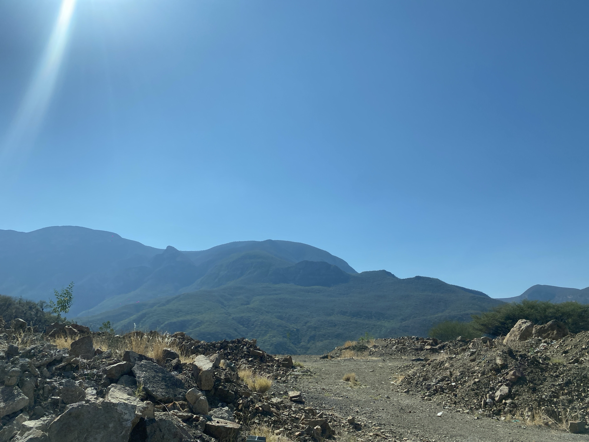 The image captures a landscape near Cerro Verde, Mexico. The foreground consists of a dirt path flanked by rocky terrain. The rocks are light gray and brown. Sparse vegetation, including dry grasses and small shrubs, is scattered amidst the rocks. In the mid-ground, the scene transitions to a view of distant, hazy mountains with dense green vegetation. The sky is a clear, light blue. A bright glare from the sun is visible in the upper-left corner of the frame. There are no people visible in the image. The environment is arid.