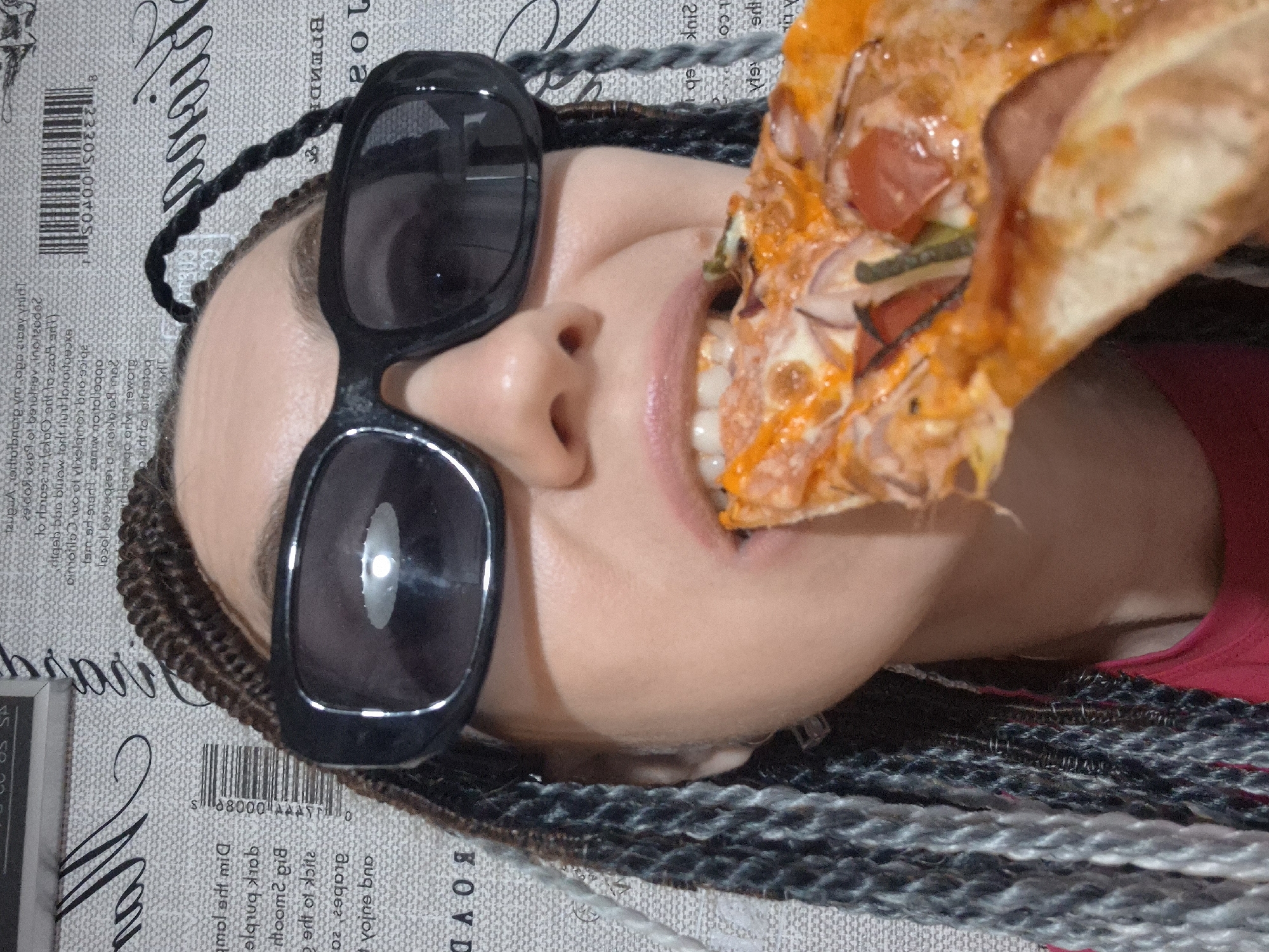 This is a close-up, vertical, overhead shot of a person eating pizza. The person, whose face is mostly obscured by sunglasses and a slice of pizza, has dark braided hair that trails down the sides of their head and across the background. The background is a light-colored, textured surface with what appears to be text and a barcode printed on it. The pizza slice is large and appears to have cheese, tomato, onion, and possibly sausage or another meat topping. The person's mouth is open, about to take a bite of the pizza. The lighting suggests an indoor setting, as there are no strong shadows or indications of natural light. The overall impression is casual and perhaps a bit messy, with the pizza being the clear focal point of the activity. Visible text in the background includes "BOVD" and a barcode with the numbers "88000" and "8233050005".
