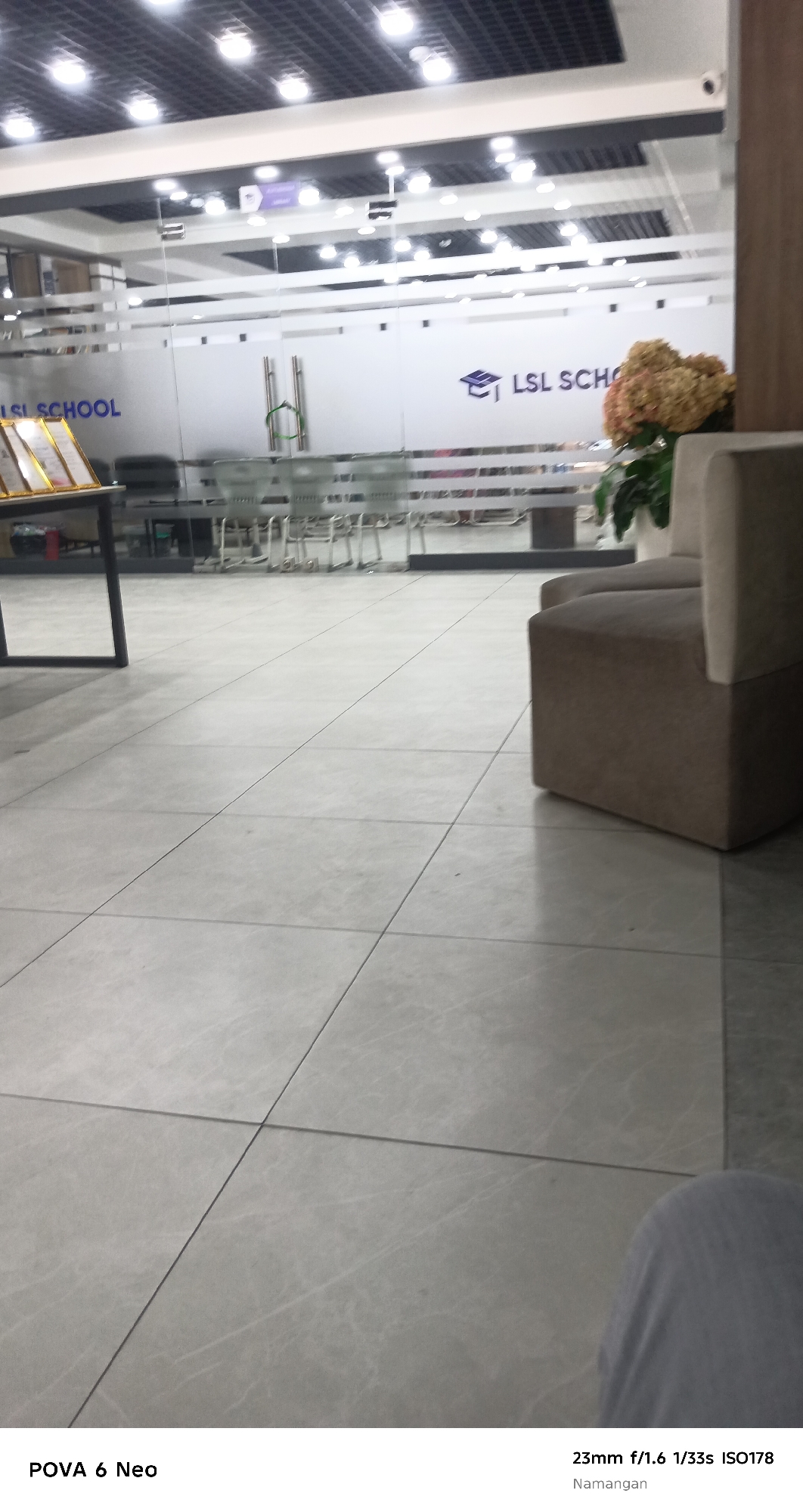 The image shows the interior of what appears to be a school or educational institution, likely located in Namangan City, Uzbekistan. The foreground is dominated by a tiled floor. In the background, there are glass doors with frosted lettering that reads "LSL SCHOOL." Beyond the doors, a row of chairs and tables are visible, suggesting a seating or study area. To the right of the entrance, a beige-colored sofa or bench is positioned. A potted plant with dried flowers sits near the sofa. The ceiling features a grid of lights. The bottom of the image displays text identifying the device and camera settings: "POVA 6 Neo" and "23mm f/1.6 1/33s ISO178". The lighting suggests an indoor environment during the day. There are no people clearly visible in the frame.