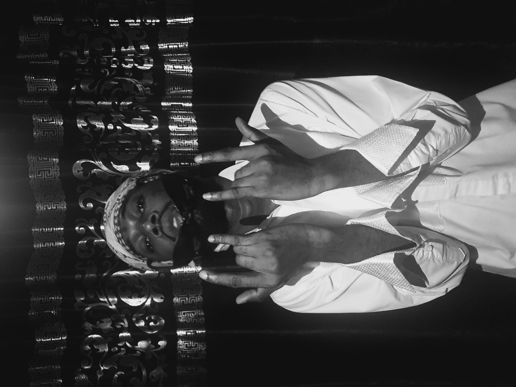 A black and white photograph depicts a man lying down in front of what appears to be a patterned wall or surface. The man wears a head covering and a light-colored shirt, possibly white or very light gray. He is making hand gestures towards the viewer with both hands. The setting and the man's clothing are generic enough that the location in Lokoja, Nigeria cannot be determined from the visual information available. There is no visible geographical or identifying background.