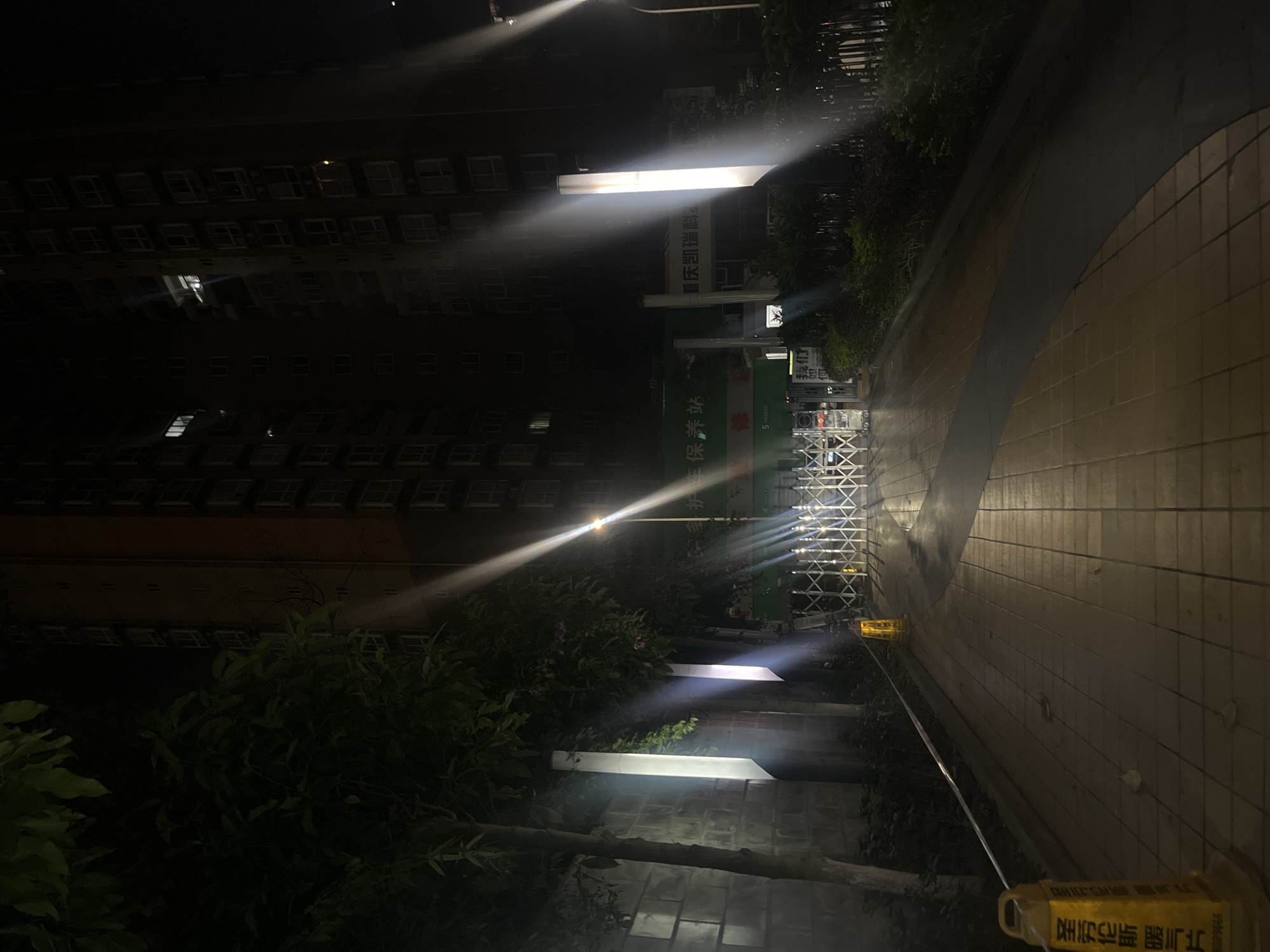 This nighttime image captures the entrance to a building complex or facility in Jiulongpo District, Chongqing, China. The scene is illuminated by multiple artificial light sources, creating bright streaks and reflections on a potentially damp paved surface.
In the background, two multi-story residential or commercial buildings stand tall, with numerous windows, some of which are illuminated. The building on the left appears darker, while the one on the right exhibits a brownish-orange hue. Dense green foliage, consisting of trees and bushes, occupies the left and central midground, partially obscuring the buildings.
The main subject in the midground is a white or silver accordion-style retractable gate, blocking access to a paved driveway. Behind this gate, on a green backdrop, two signs are visible. The left sign displays Chinese characters "养护站" (yǎnghù zhàn - maintenance station or nursing station). The right sign reads "重庆凯瑞科" (Chóngqìng Kǎiruì Kē - Chongqing Kairui Science/Technology/Department), explicitly confirming the location in Chongqing.
The paved entrance path consists of light-colored square tiles with a darker, possibly asphalt, strip running through it. Two yellow plastic warning cones are present on the path. One in the left foreground features Chinese characters "圣劳伦斯暖气片" (Shèng Láoluènsī Nuǎnqì Piàn - Saint Lawrence Radiator) and "地滑请小心" (Dì huá qǐng xiǎoxīn - Slippery ground, please be careful). Another similar cone is located near the gate on the right.
Lighting in the scene is provided by two tall, vertical white light fixtures on the left side of the entrance, and a single streetlamp on the far right. These lights cast strong, bright beams and illuminate the immediate area, highlighting moisture on the pavement. No individuals are visible in the image, suggesting the area is deserted at the time of capture.