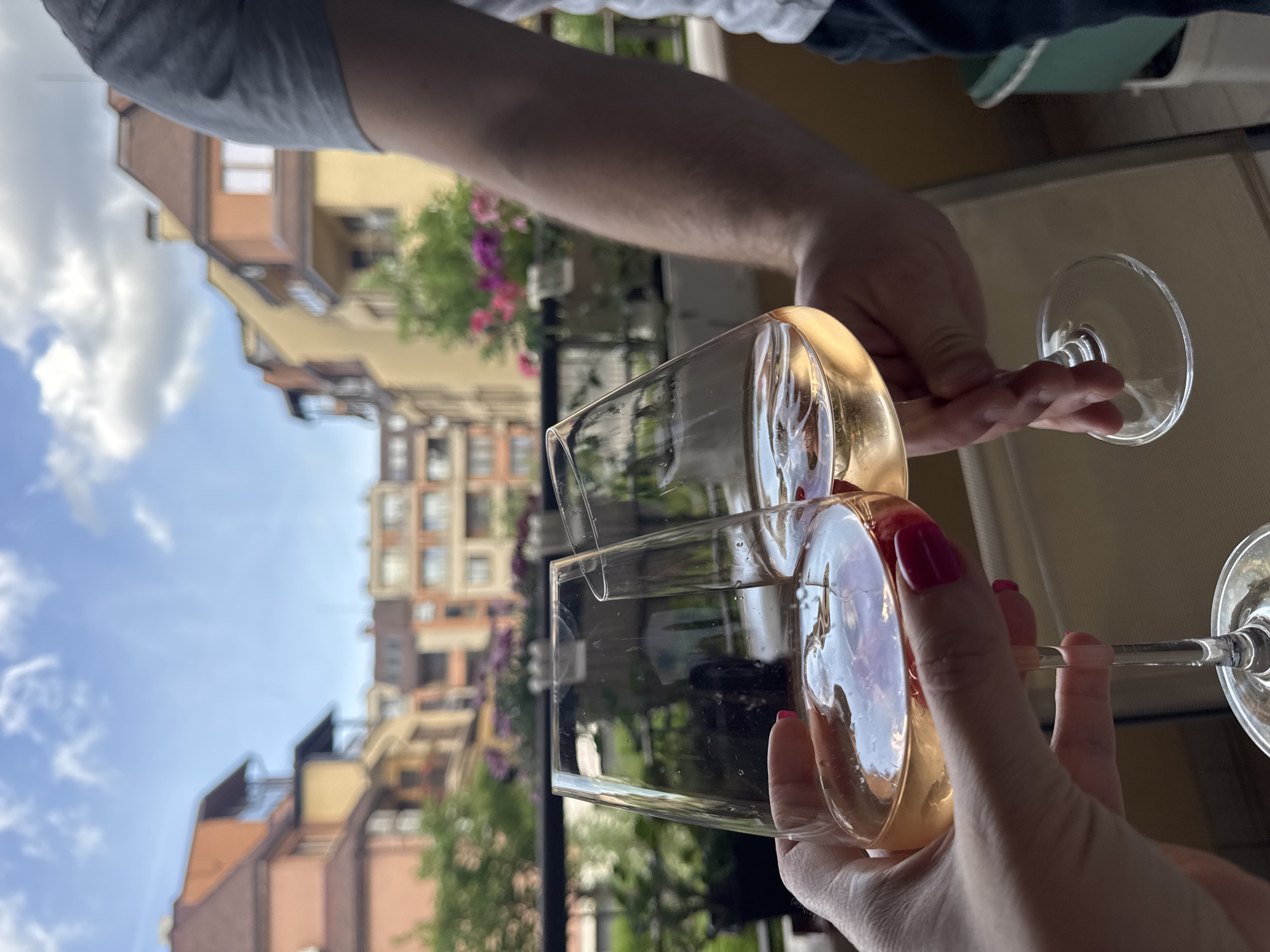 Two individuals are toasting with glasses of white wine, the glasses nearly touching. The individual on the left has pale skin and is wearing a grey t-shirt. The individual on the right is holding a round wine glass and has red nail polish. The glasses are transparent, revealing the wine inside and refracting the scene behind.
In the background, there is a view of apartment buildings. The architectural style suggests a location in Sofia, Bulgaria, specifically in the vicinity of Boyana district. These buildings are multi-storied, with various architectural details and colors. The sky is visible in the upper left corner of the image. There are greenery and flowers visible on the balconies of the buildings. The scene appears to be taken on a balcony.