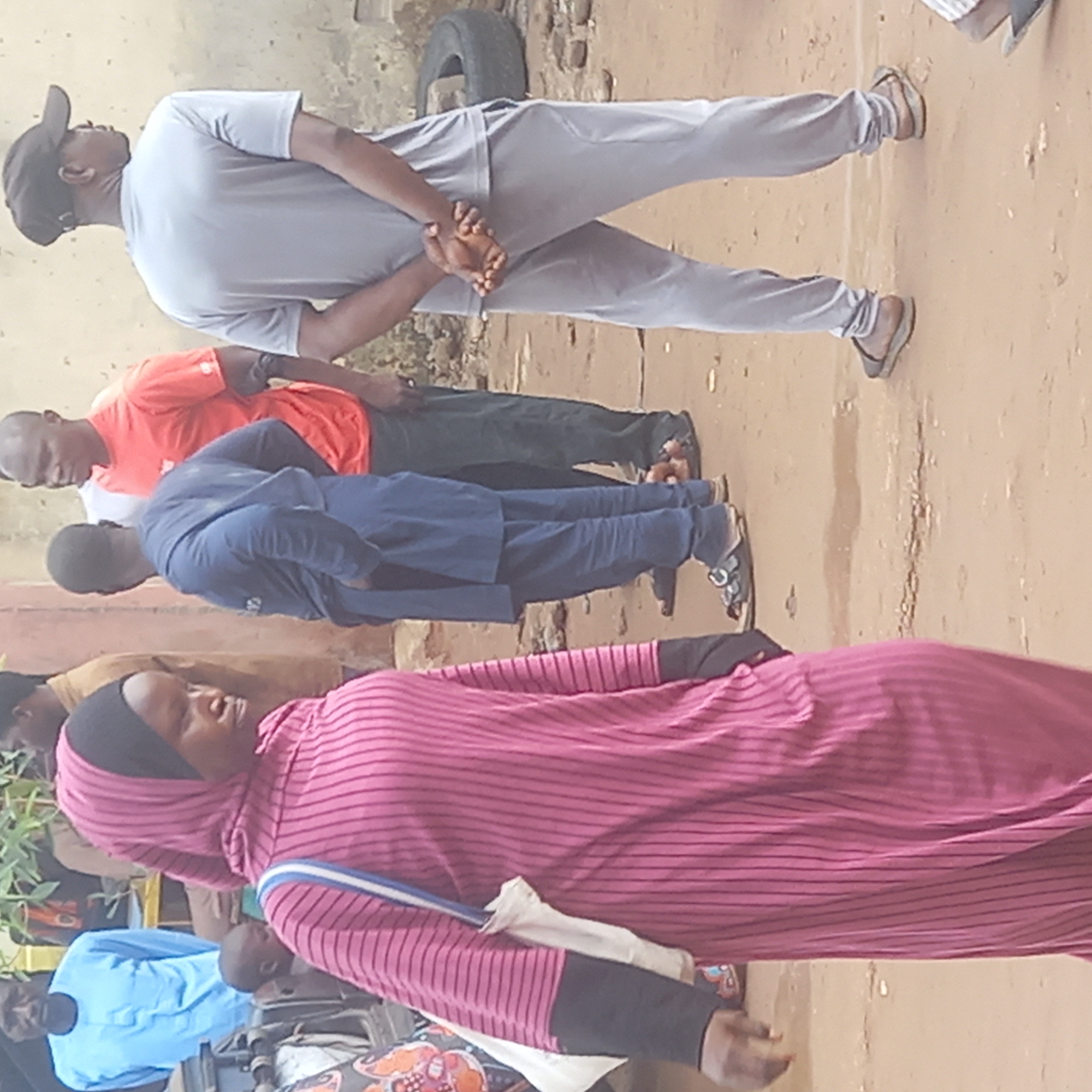 The image shows a group of people standing outdoors, seemingly on a dirt surface. From the orientation of the image, the people are standing in a line from the foreground to the background. The person closest to the viewer is wearing a pink striped dress with a hijab. Behind her is a man in a blue outfit. Further back, there's a man in an orange shirt and a person in a white shirt, and finally a man in a grey sweatsuit with a baseball cap. The individuals are all looking forward. In the background, a wall is visible, and a tire is leaning against it. The location is assumed to be Dadi Riba, Nigeria, based on the prompt.