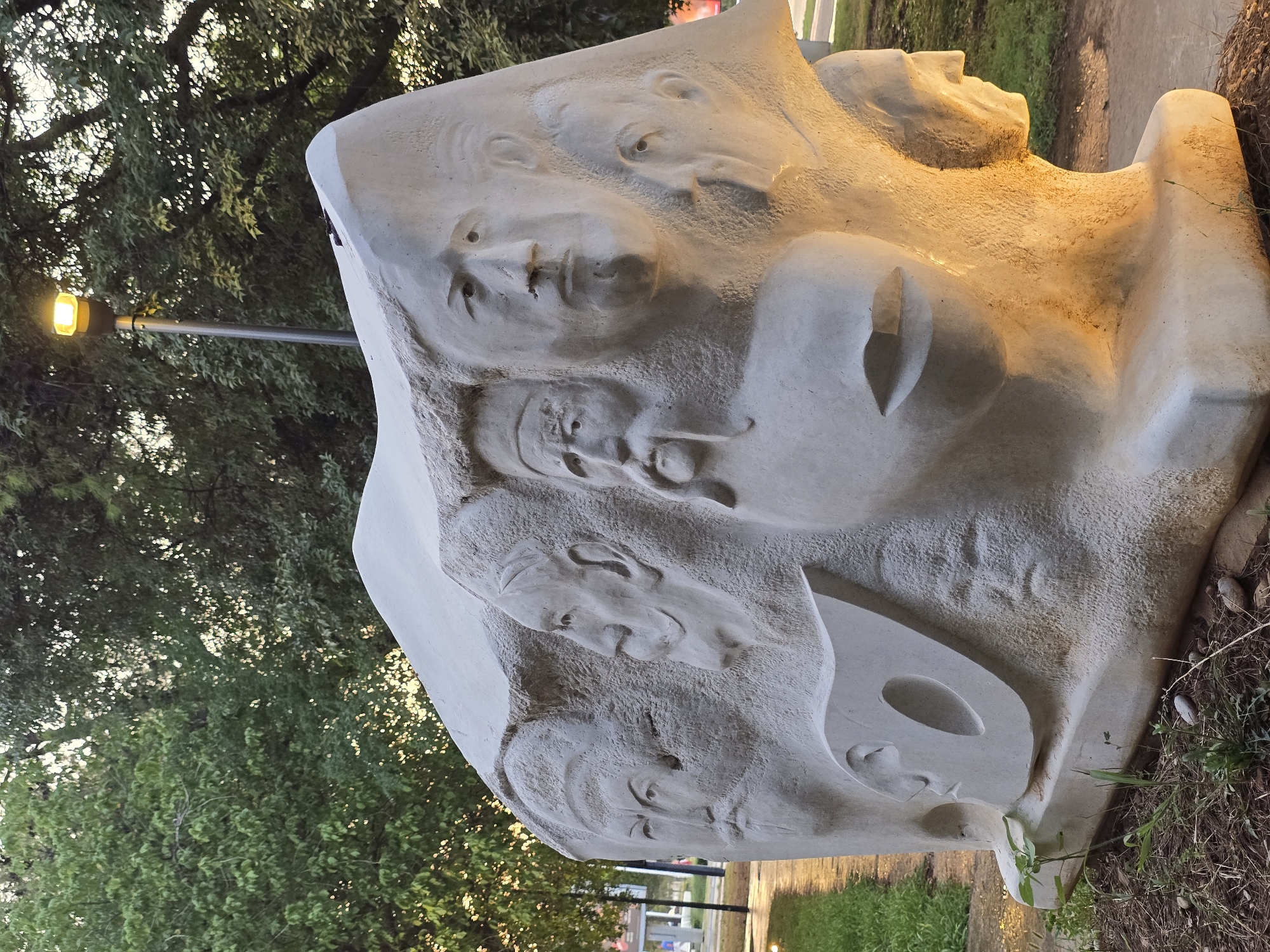 The image depicts a large, abstract, light-colored stone sculpture featuring multiple carved human faces in profile and full view, some with beards and mustaches. The sculpture is situated outdoors, possibly in a park or public space, with trees and greenery visible in the background. A street lamp with a yellow light is positioned behind and to the right of the sculpture. The ground around the sculpture is covered in mulch and some sparse vegetation. The lighting suggests it is either dusk or dawn, with the street lamp providing artificial illumination that casts a warm glow on the right side of the sculpture. The sculpture appears to be a significant public art installation. It is located in Podgorica, Montenegro, specifically in the park area of Kruševac.