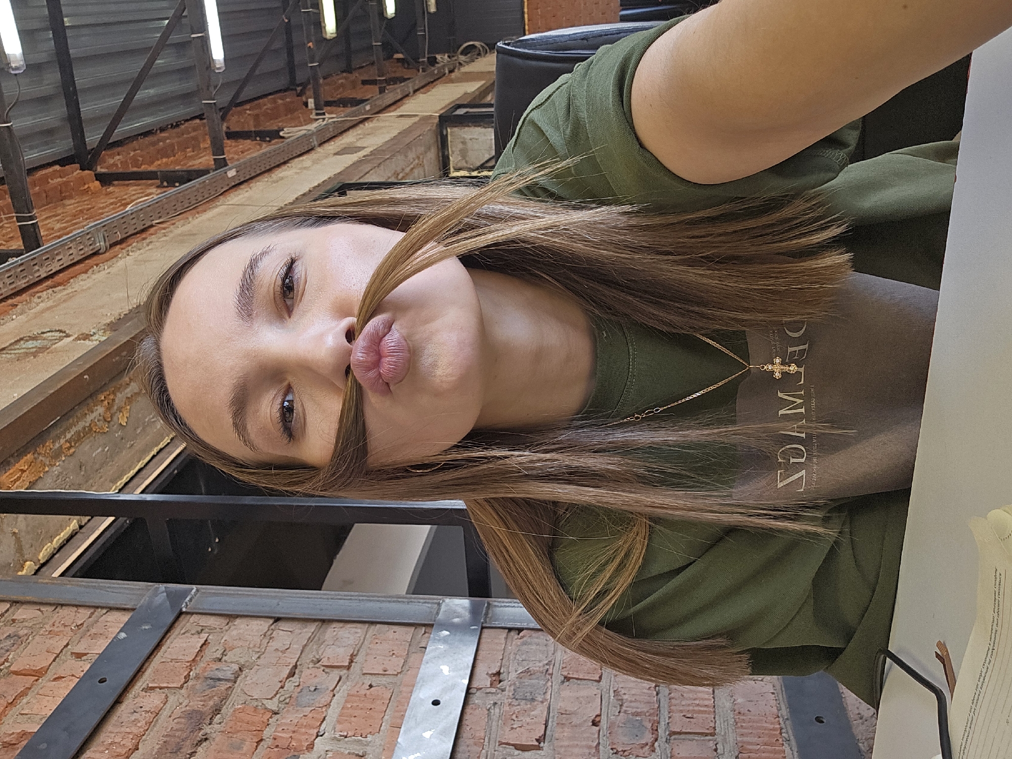 A woman is taking a selfie in what appears to be an industrial or loft-style space. The woman has long, light brown hair, part of which is styled to resemble a mustache over her pouting lips. She is wearing a green t-shirt and a gold necklace with a cross pendant. The background features exposed brick walls, metal beams, and industrial lighting fixtures. There is a black chair visible and some papers on a table. Based on the information given, the location is in the city of Chaikovsky, Russia.