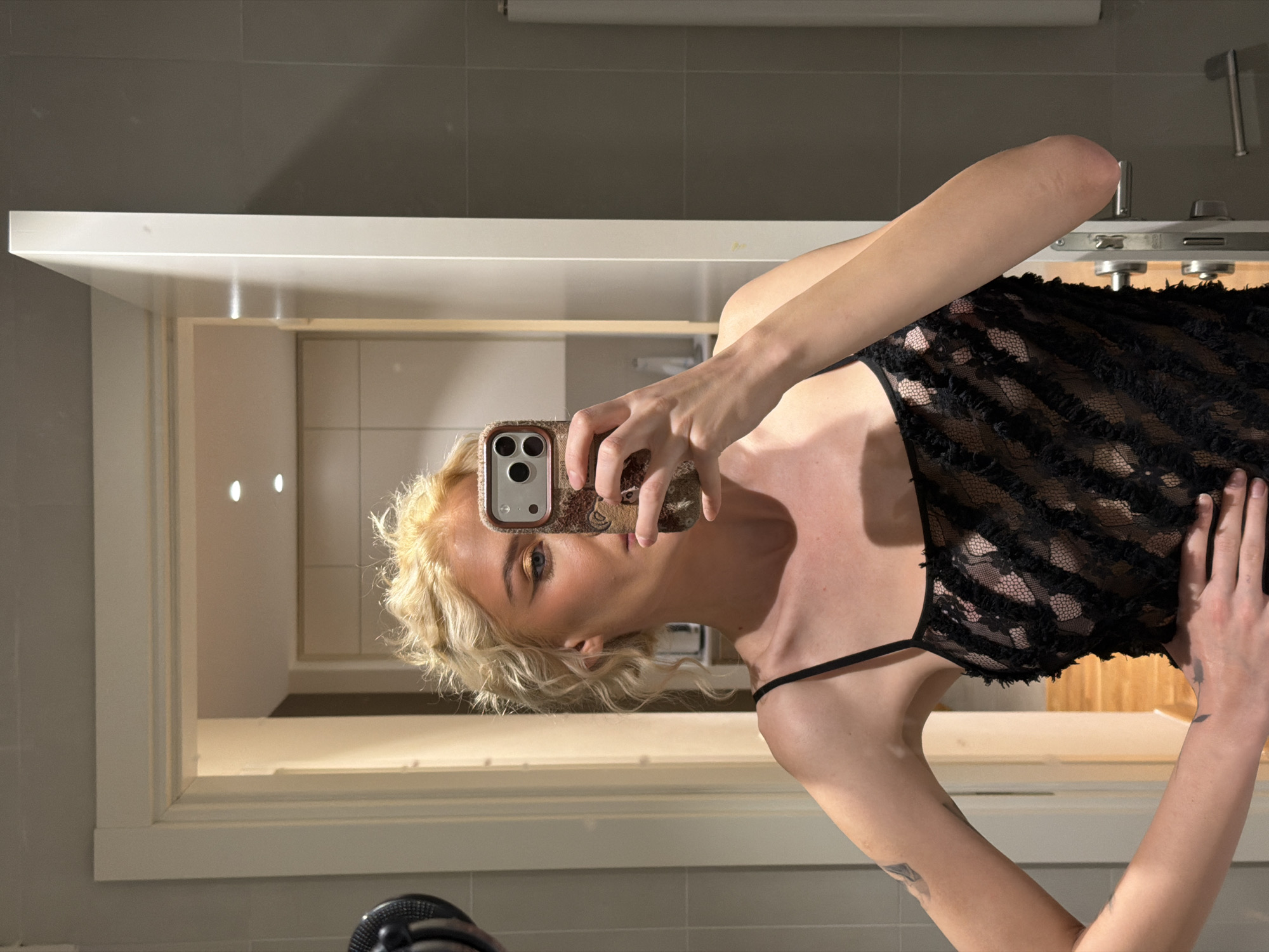 A woman with short blonde hair takes a selfie in a bathroom mirror. She is wearing a black lace top with spaghetti straps. She is holding a phone with a camouflage case. The bathroom has grey and white tiles, a white sink and countertop, and a silver faucet. The background suggests a modern or renovated interior. The photograph's location cannot be determined from the image.