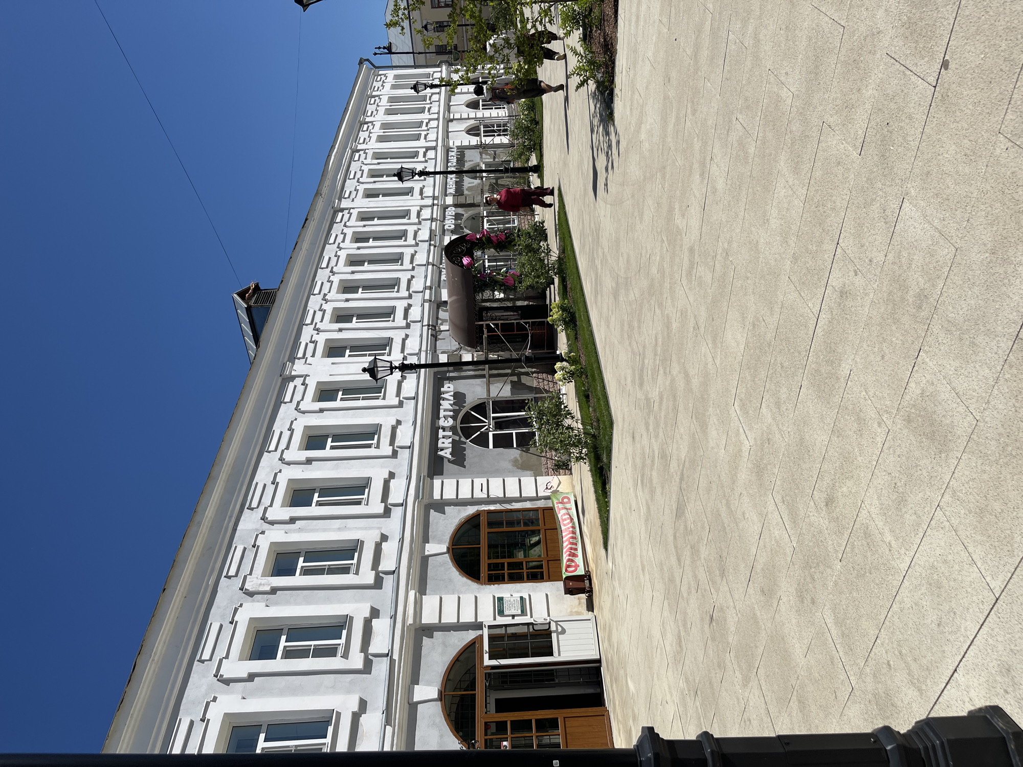 The image shows a street scene in Novo-Nikolskoye, Russia, on a bright, sunny day with a clear blue sky. The main focus is a long, two-story building with a light-colored facade, featuring numerous windows with decorative white trim. The ground floor of the building contains several storefronts. One storefront, labeled "ART стиль", has a large arched window. To its left, there are two entrances with arched windows above them. A green banner with red Cyrillic text is displayed on the sidewalk in front of one of these entrances.
Several people are walking on the wide, paved pedestrian area in front of the building. On the right side of the image, two individuals are walking away from the camera. To the left of them, a person in a dark red outfit is walking towards the camera. The pedestrian area is paved with large, light-colored rectangular tiles arranged in a staggered pattern. Lampposts are visible along the sidewalk. There are also some bushes and small trees planted in beds along the building's facade and the pedestrian area. The overall scene depicts a clean and well-maintained urban environment.
