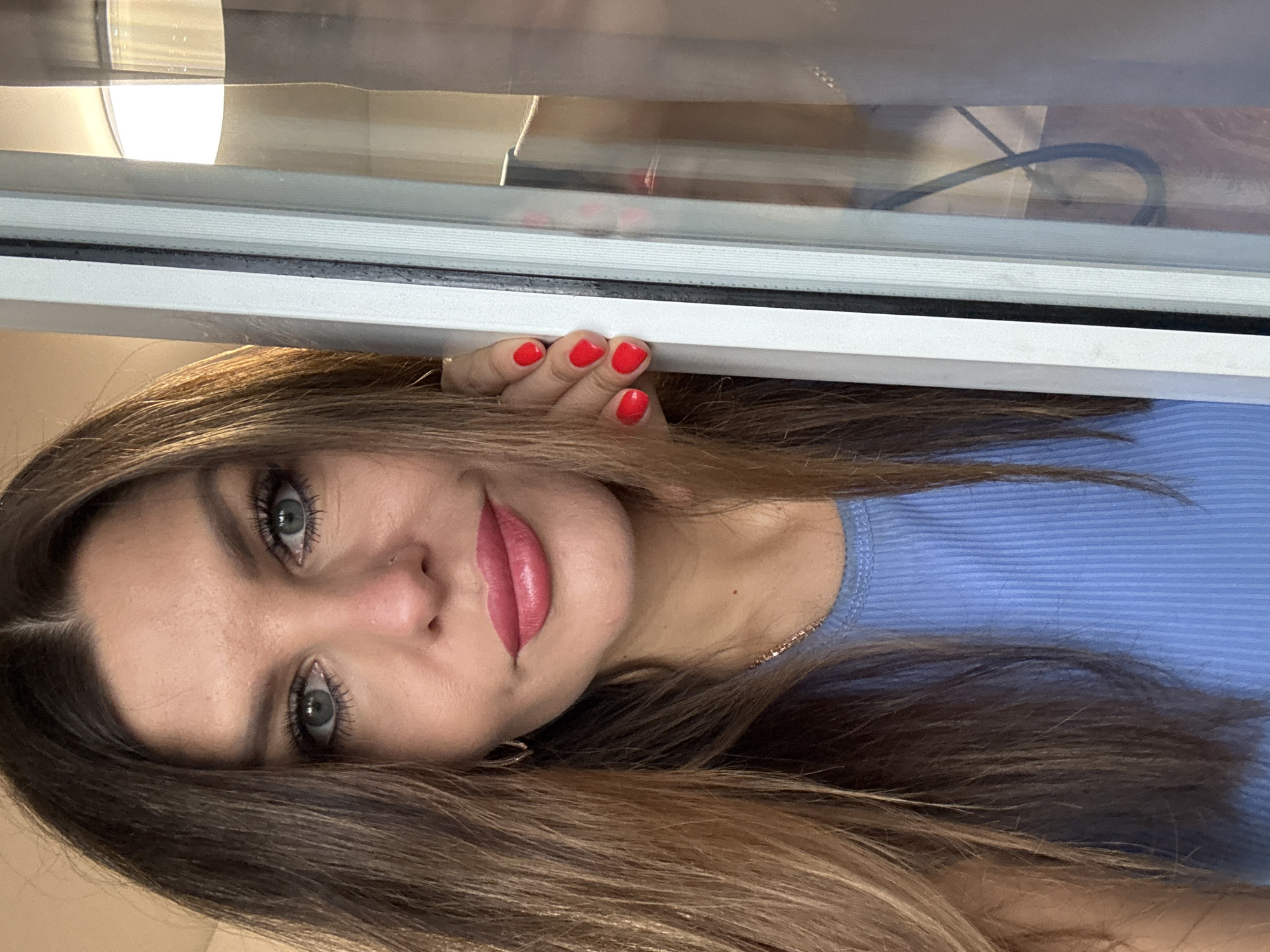 A woman with light brown, layered hair, green eyes, and red lipstick is positioned under a white window frame, holding onto the frame with her right hand. Her nails are painted red. She is wearing a light blue ribbed top. The background visible through the window is blurry but appears to be a neutral-colored interior. The image is taken at an angle. The image contains no information to identify that it was taken in Chaikovsky, Russia.