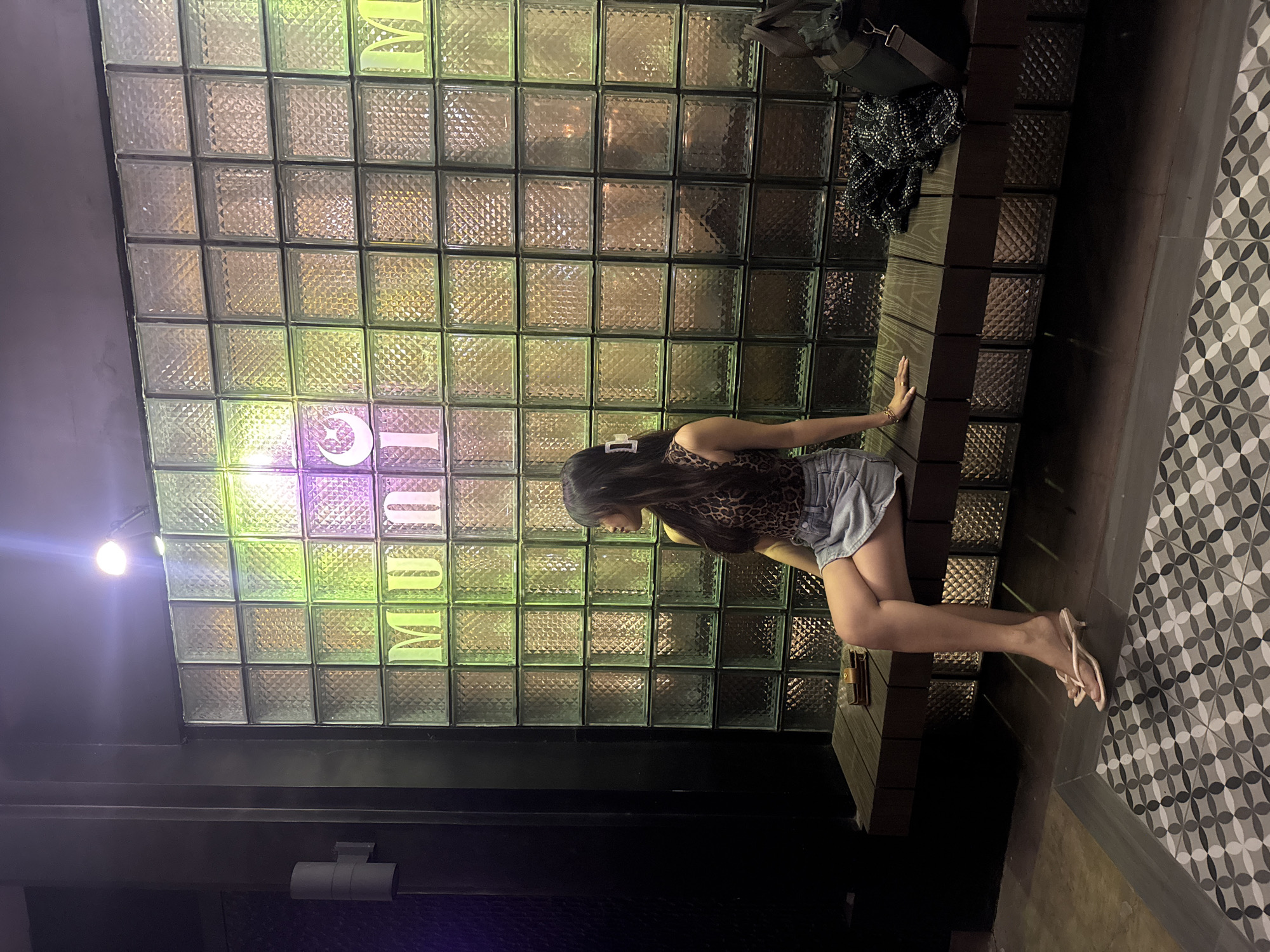 A woman with long dark hair, wearing a leopard print halter top and a denim skirt, sits on a wooden bench in front of a wall made of textured glass blocks. The glass block wall is illuminated by neon lights that cast colorful hues of green, yellow, and purple. The word "MUNICIPAL" is spelled out in capital letters, with the letters appearing in white and illuminated. To the right of the woman on the bench are a black handbag and a tweed jacket. The floor in front of the bench has a patterned tile design. The scene appears to be an indoor location, possibly a lounge or entertainment venue. The woman is looking to her right, away from the camera.