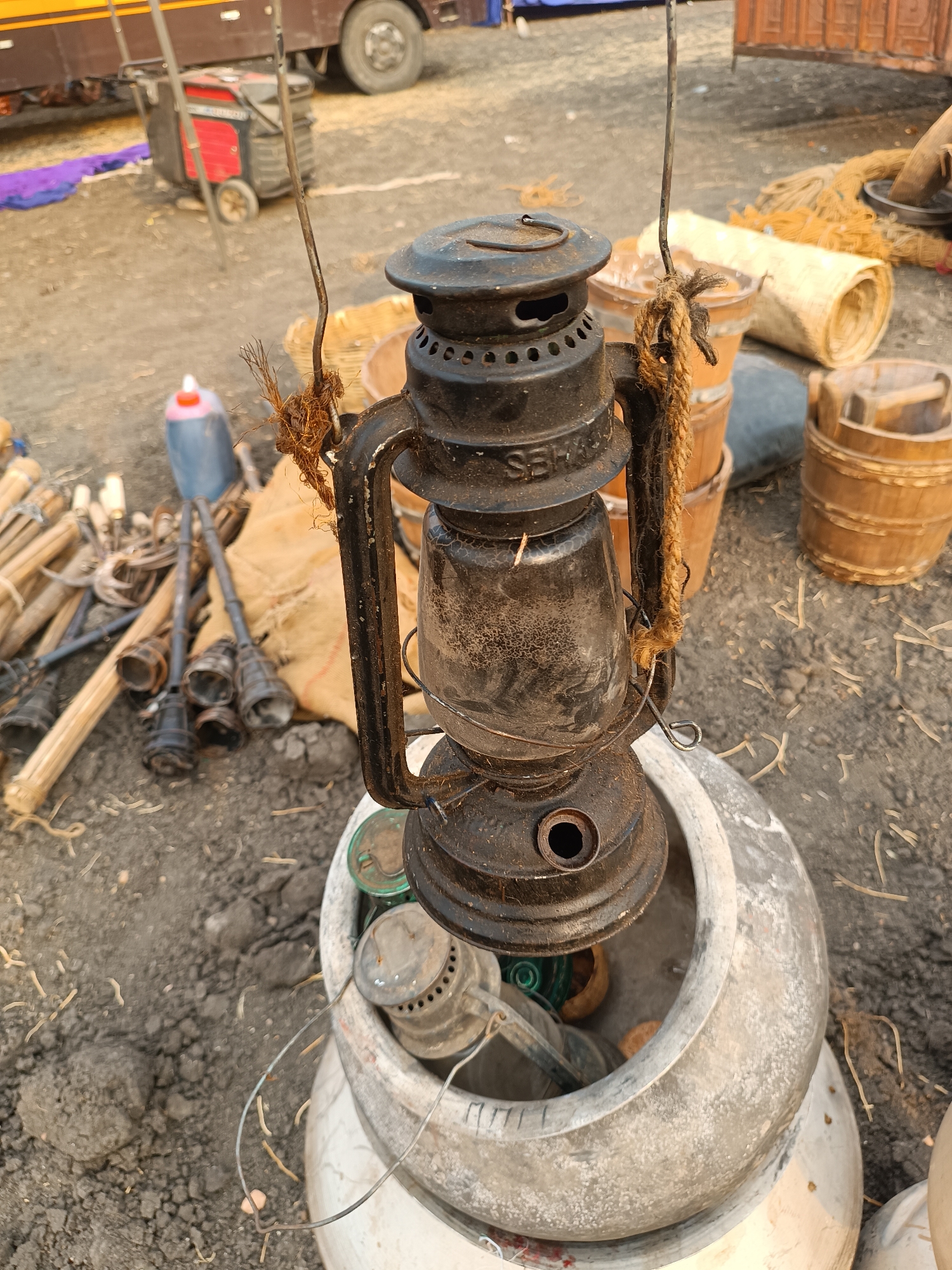 The image depicts an outdoor scene in Kottalapalli, India, on dry, uneven dark earth mixed with sparse organic debris. A weathered, dark-colored traditional-style kerosene lantern, inscribed 'SEH' on its side, is suspended by thin metal wires and a knotted brown rope. The lower section of the lantern rests within a large, light-grey, textured ceramic pot that also contains smaller metallic objects.
To the left of the central arrangement, a collection of long wooden poles or bamboo sticks lies alongside several dark-colored, flared tubular objects. A blue plastic container with a red cap is also visible. Further back, a red and black portable generator on wheels is positioned near a folded piece of purple fabric, beneath the visible section of a large brown vehicle.
To the right of the central pot, multiple light brown wooden barrels and cylindrical containers are present, along with rolled straw mats and coils of light-colored rope or twine. A brown rectangular structure or part of another vehicle is partially visible in the upper right background. No individuals are present, and no overt actions or interactions are depicted.