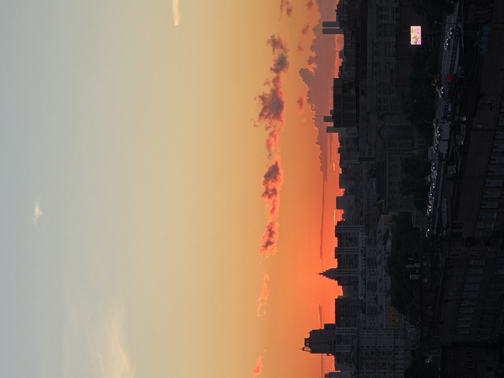 The image shows a wide shot of Moscow, Russia during sunset. The sky is a gradient of orange and yellow, with some pinkish clouds scattered across it. In the foreground, there is a dark silhouette of a cityscape, with many buildings of varying heights visible. The buildings are mostly dark and appear to be residential or office buildings. There is a highway in the midground with cars driving on it, their headlights on. In the distance, there are two tall, slender buildings that are characteristic of Moscow's skyline. The overall impression is one of a bustling city at dusk, with the warm colors of the sunset providing a dramatic backdrop.