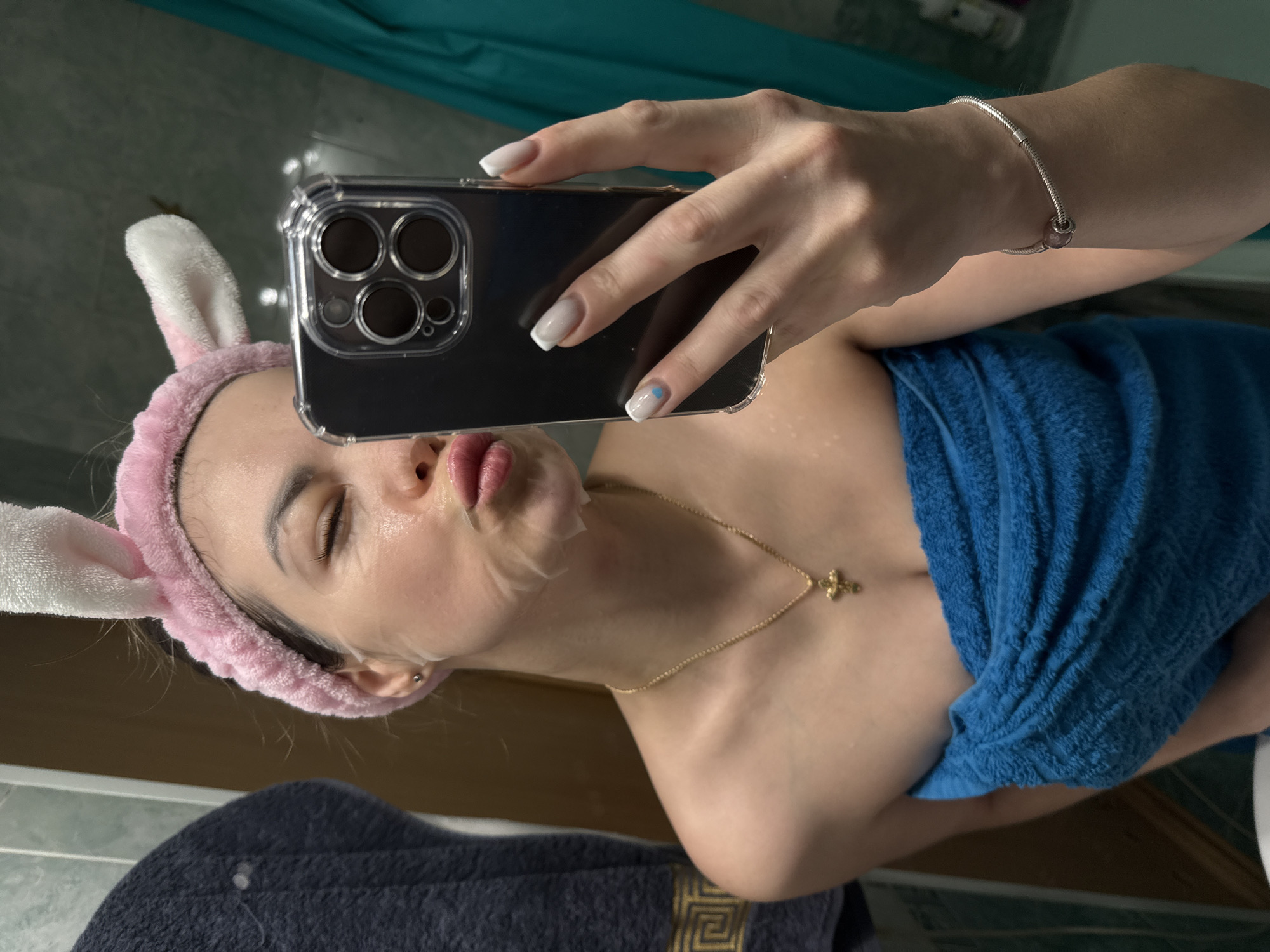 A person is taking a selfie in a bathroom. The person is wearing a pink headband with bunny ears, a gold necklace with a cross pendant, and a facial sheet mask. They are holding a smartphone up to their face, and a blue towel is draped around their torso. The person's nails are manicured with white tips, and there is a silver bracelet on their wrist. The background includes a blue shower curtain, a wooden cabinet, and a dark towel with a gold pattern. The city and location are unknown.