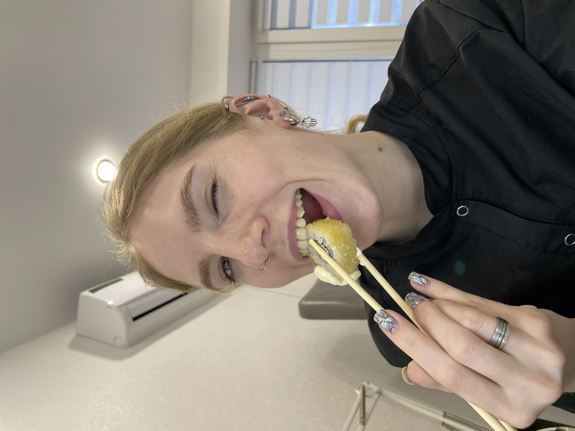 A young woman, likely in Izhevsk, Russia, is captured mid-meal in a brightly lit indoor setting. Her blonde hair is pulled back, revealing multiple ear piercings, including an industrial bar and a Hamsa hand earring on her right ear, and a nose ring. She is smiling broadly and winking with her left eye, conveying a playful and joyful expression as she brings a fried sushi roll to her mouth with wooden chopsticks. White sauce is visible on the sushi. Her fingernails are manicured with a light base and intricate silver or white floral designs. She wears a multi-band silver ring on her left ring finger and a black garment, possibly a uniform or casual top. In the background, a white air conditioner unit is mounted on a wall, and a circular ceiling light fixture illuminates the space. A window with vertical blinds is also visible. No text is discernible in the image.