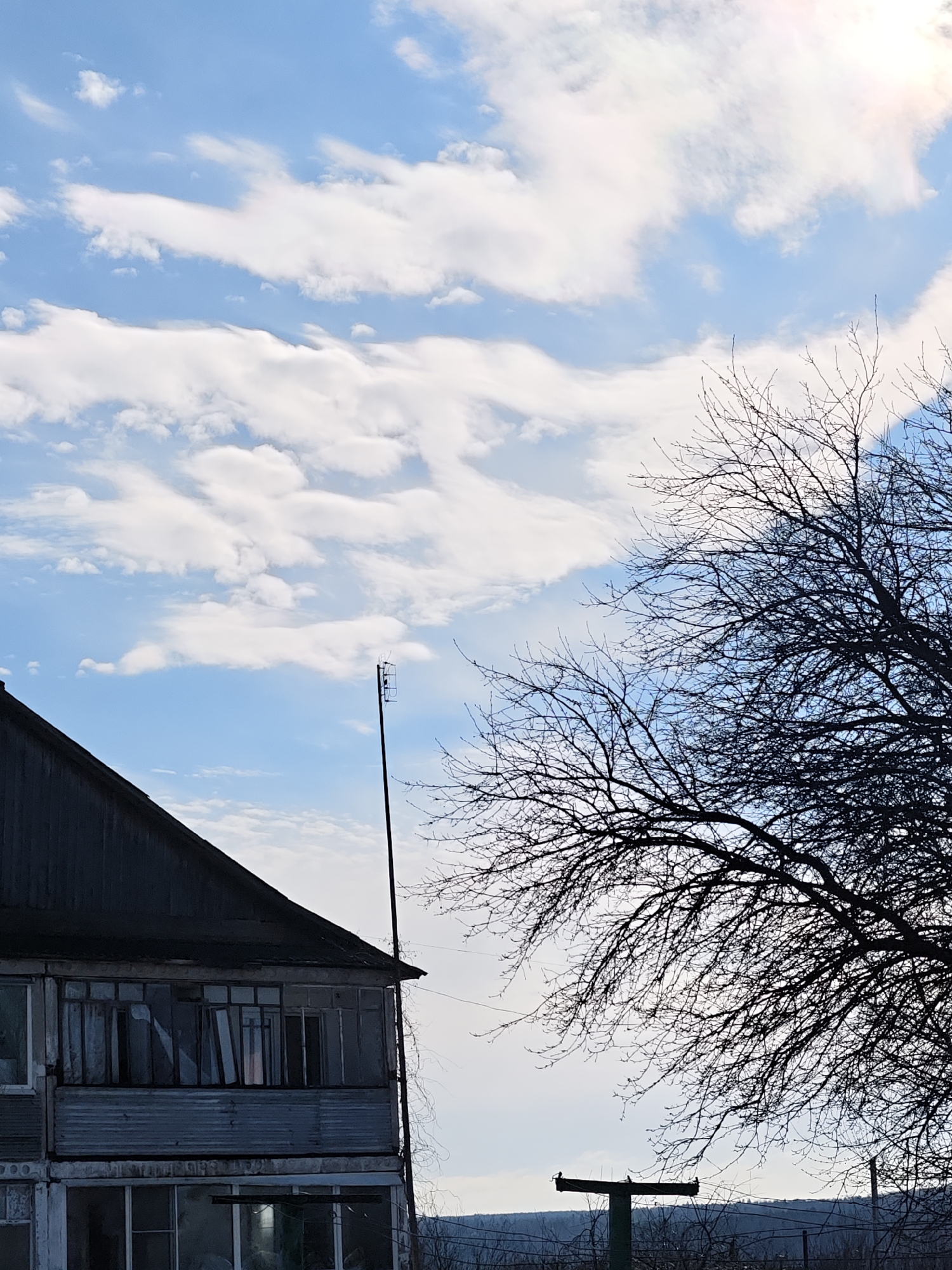 The image shows a partly cloudy sky above a two-story building with a dilapidated exterior in the city of Уральское, Russia. A tall antenna is affixed to the building's side. A large, bare tree occupies the right side of the frame. In the background, a distant treeline is visible.