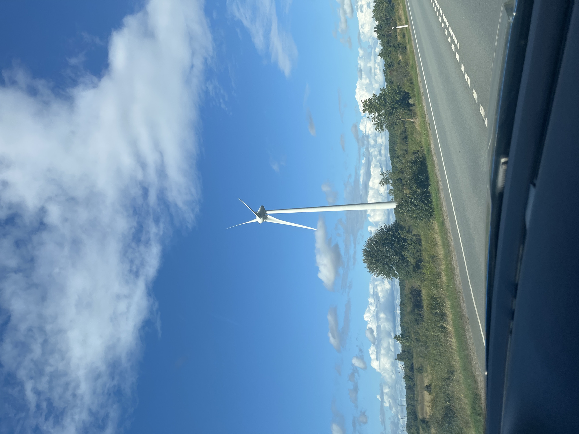 This is an eye-level, outdoor, medium shot capturing a vast blue sky filled with scattered white clouds. In the mid-ground, a single white wind turbine stands tall, its blades angled upwards. To the right of the turbine, a paved road with white lane markings curves away into the distance. The landscape flanking the road is a mix of green fields and trees, with more wind turbines visible in the far distance, suggesting a wind farm. The image appears to be taken from a vehicle, as the dashboard is visible in the foreground, partially obscuring the lower part of the scene. The overall atmosphere is bright and clear, indicative of daytime. The location is identified as Virtsu, Estonia.