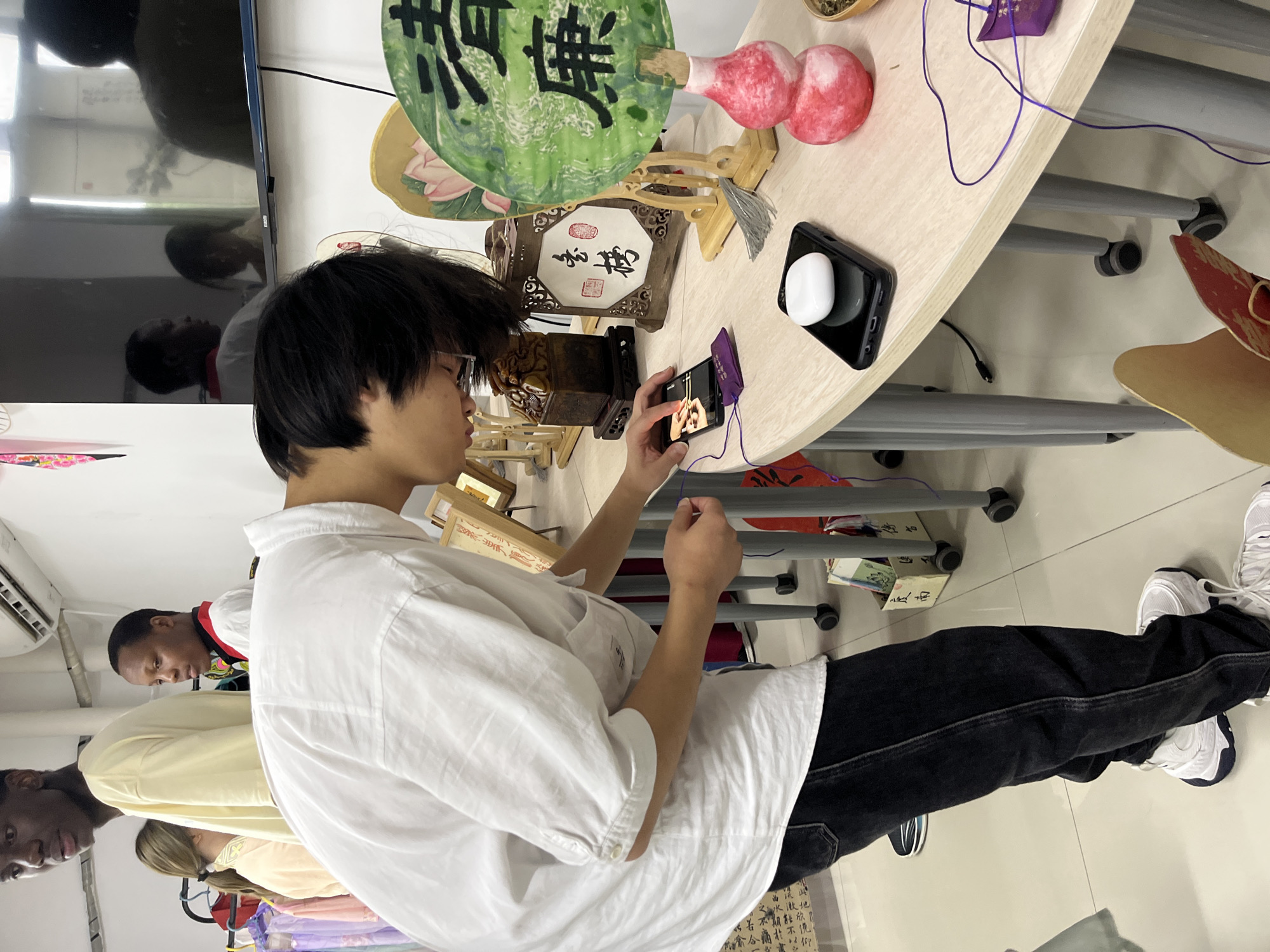 A young person is standing, wearing a white shirt and black pants, and holding a cell phone in their hands, appearing to be filming an arrangement of traditional Chinese artifacts. The objects include what appears to be a fan with Chinese writing, a wooden lantern, a gourd-shaped object, and other decorative pieces on a light-colored table. There are two other people standing behind the subject. The background includes an air conditioner, a television screen with a reflection, and what looks like a classroom or studio setting. The Chinese characters suggest this is in Jiangjin, China.