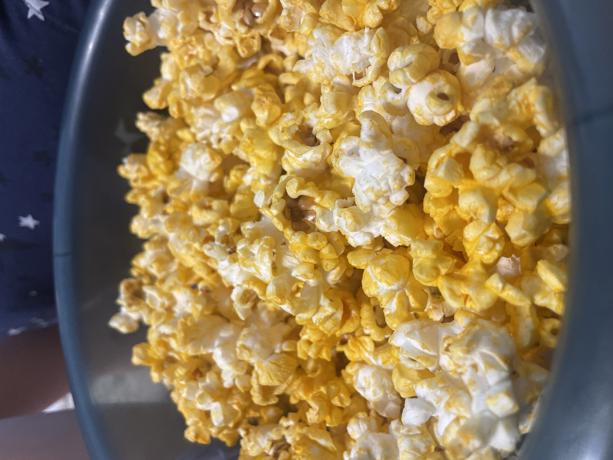 A close-up shot from above captures a blue bowl filled with popcorn, some of which is coated in yellow flavoring. The background shows a partial view of a navy blue shirt with white stars. There's no evidence of this image being located in Vladivostok, Russia.
