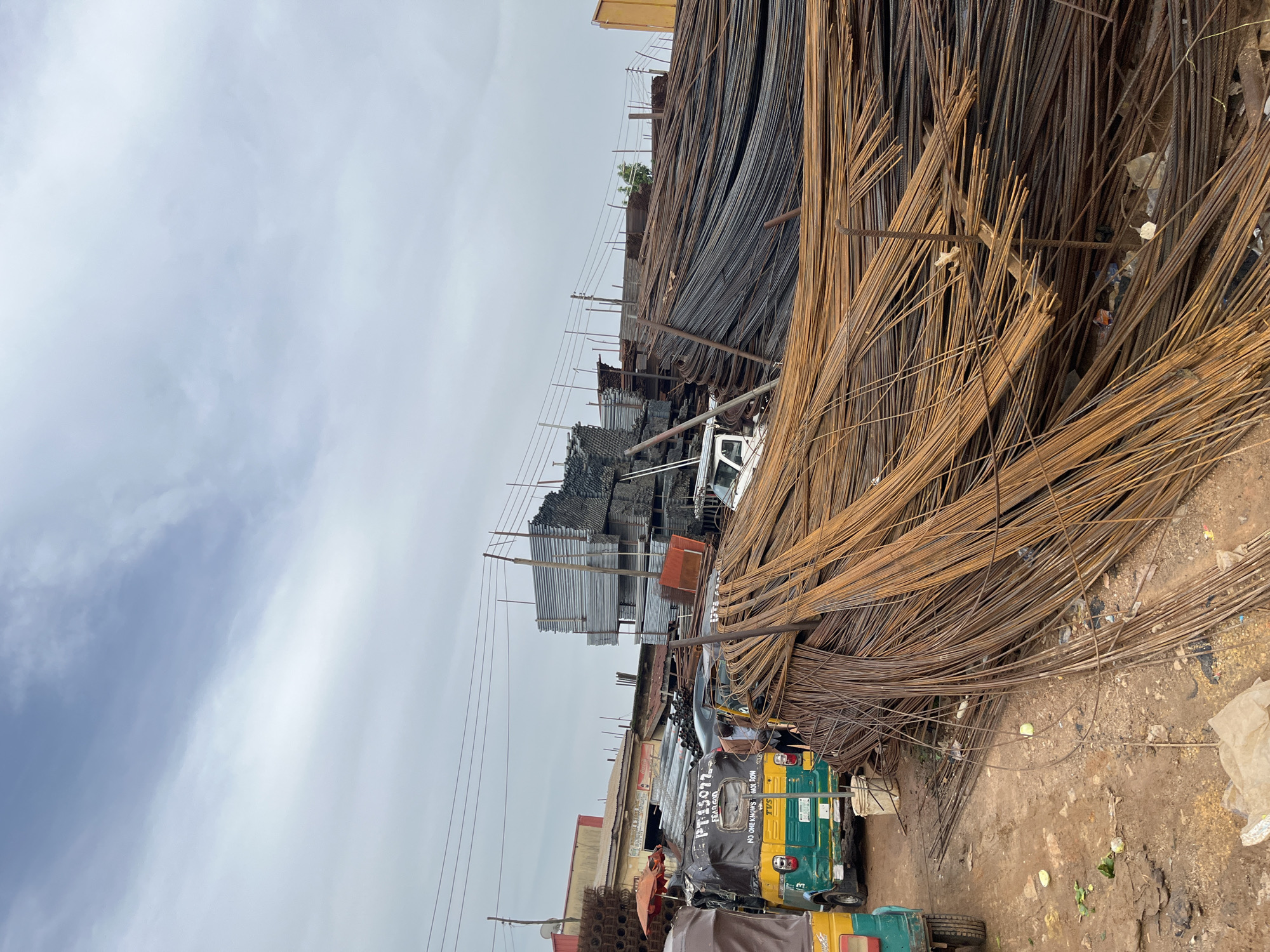 This media file captures an outdoor scene in Gura Top, Nigeria, likely a marketplace or a storage yard for building materials. The dominant subjects are large piles of steel rebar, some rusted and a darker, newer batch. In the foreground, a three-wheeled auto-rickshaw, painted yellow and green, is parked. The auto-rickshaw has text on its side, including "PT 15077", "FEAR GOD", and "NO ONE KNOWS, KNOW KNOW".
Other objects visible include stacks of corrugated metal sheets and various metal rods, suggesting a construction or metalworking vicinity. A few people are present, including one driver or operator of the auto-rickshaw. The scene is bustling with activity, indicated by the presence of the vehicle and the implication of commerce or trade in building materials.
The weather appears overcast with a cloudy sky, suggesting it might be a humid day, common in Nigeria. The ground is dirt and dusty, typical of outdoor markets and storage areas. The overall impression is one of industrial activity and a lively, perhaps somewhat chaotic, marketplace environment. The time of day is indeterminate but appears to be daylight hours.