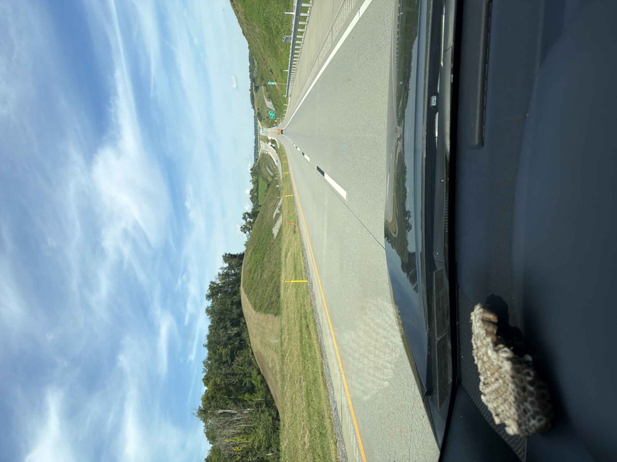 A vehicle is driving on a highway in South Fayette Township, United States of America. The view is from inside the vehicle, looking forward through the windshield. The highway is a multi-lane road with grassy shoulders and rolling hills on either side. There are trees on the hills, and a clear blue sky with wispy clouds overhead. A yellow object, possibly a vehicle or a sign, is visible in the distance on the highway. In the foreground, on the dashboard of the vehicle, is a small, honeycomb-like object.