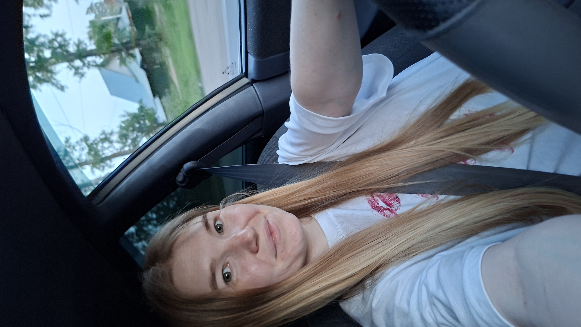 A woman with light blonde hair and fair skin is in the driver's seat of a vehicle, wearing a seatbelt and a white t-shirt with red lip prints. The car is likely moving as evidenced by the background seen through the window which features trees and a building. The location, as stated, is in the city of Chaikovsky, Russia. The woman is looking at the camera and possibly smiling. The interior of the car is primarily dark.