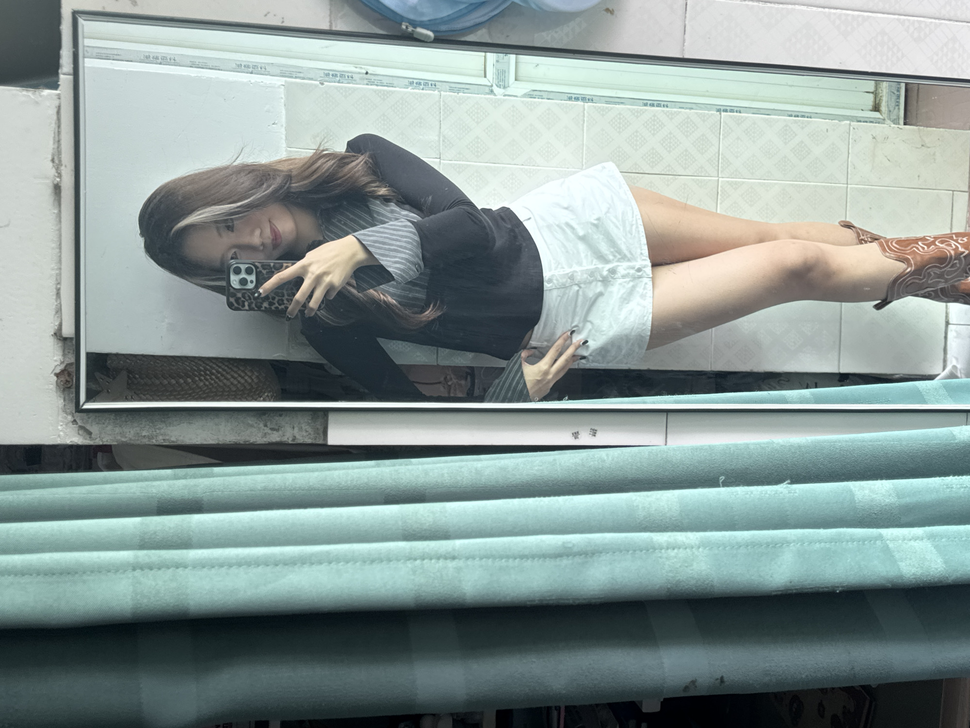 A woman is taking a mirror selfie. She is wearing a black jacket over a striped shirt, a white skirt, and brown cowboy boots. The reflection shows a tiled wall. The lower part of the image shows a set of teal-colored curtains. The location is Nan'an District, China, based on the writing on the upper window frame.