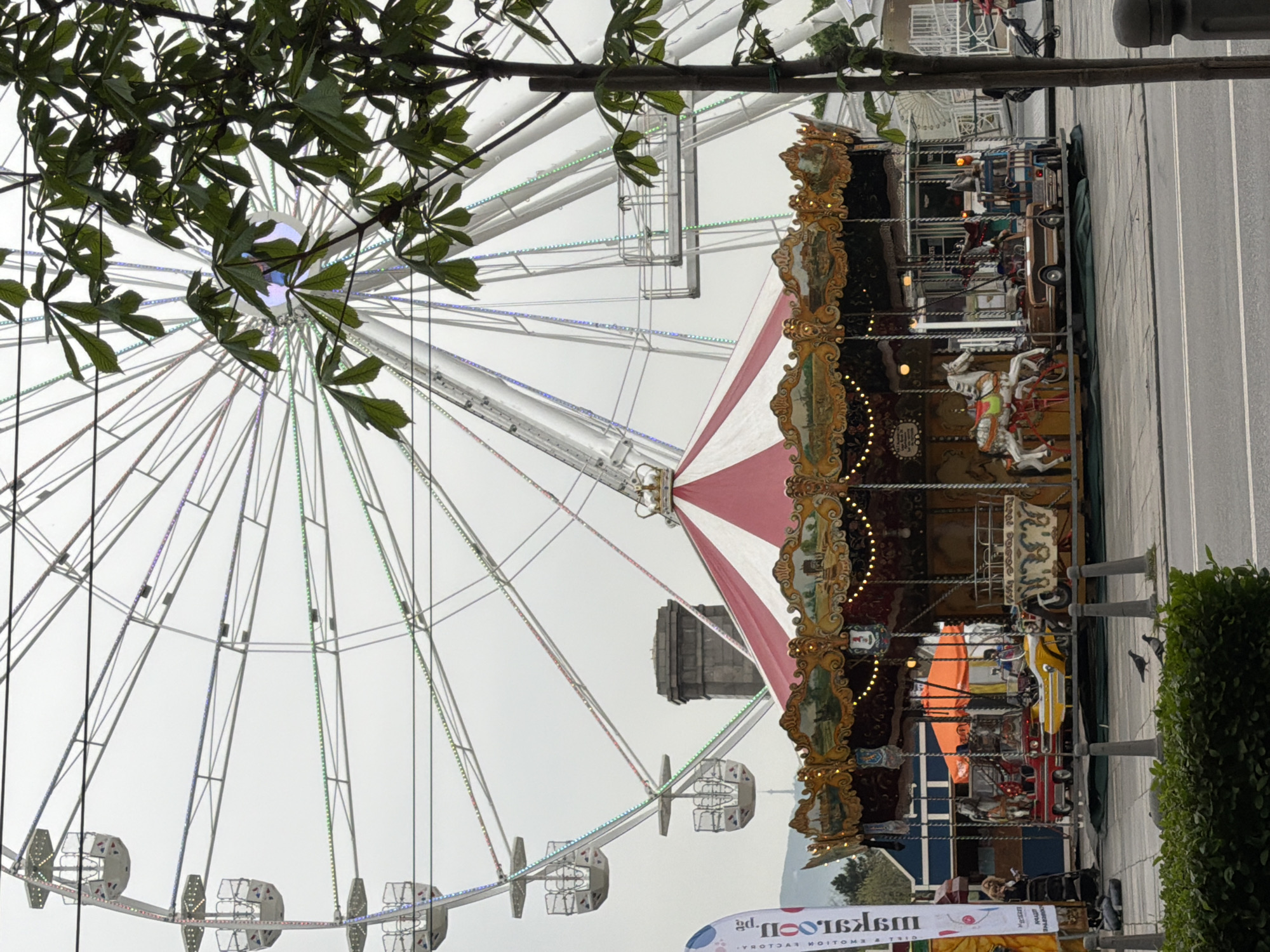 The image shows a carousel and Ferris wheel in Sofia, Bulgaria. The carousel has ornate gold decorations and painted horses. The Ferris wheel is white with colored lights and triangular passenger cars. A red and white striped tent-like structure is centrally located in the composition. A stone building can be seen behind the carnival rides. A hedge and a row of bollards are situated next to the carousel. Part of a banner is visible in the foreground. The sky is overcast, and a tree with green leaves occupies the upper left portion of the frame.