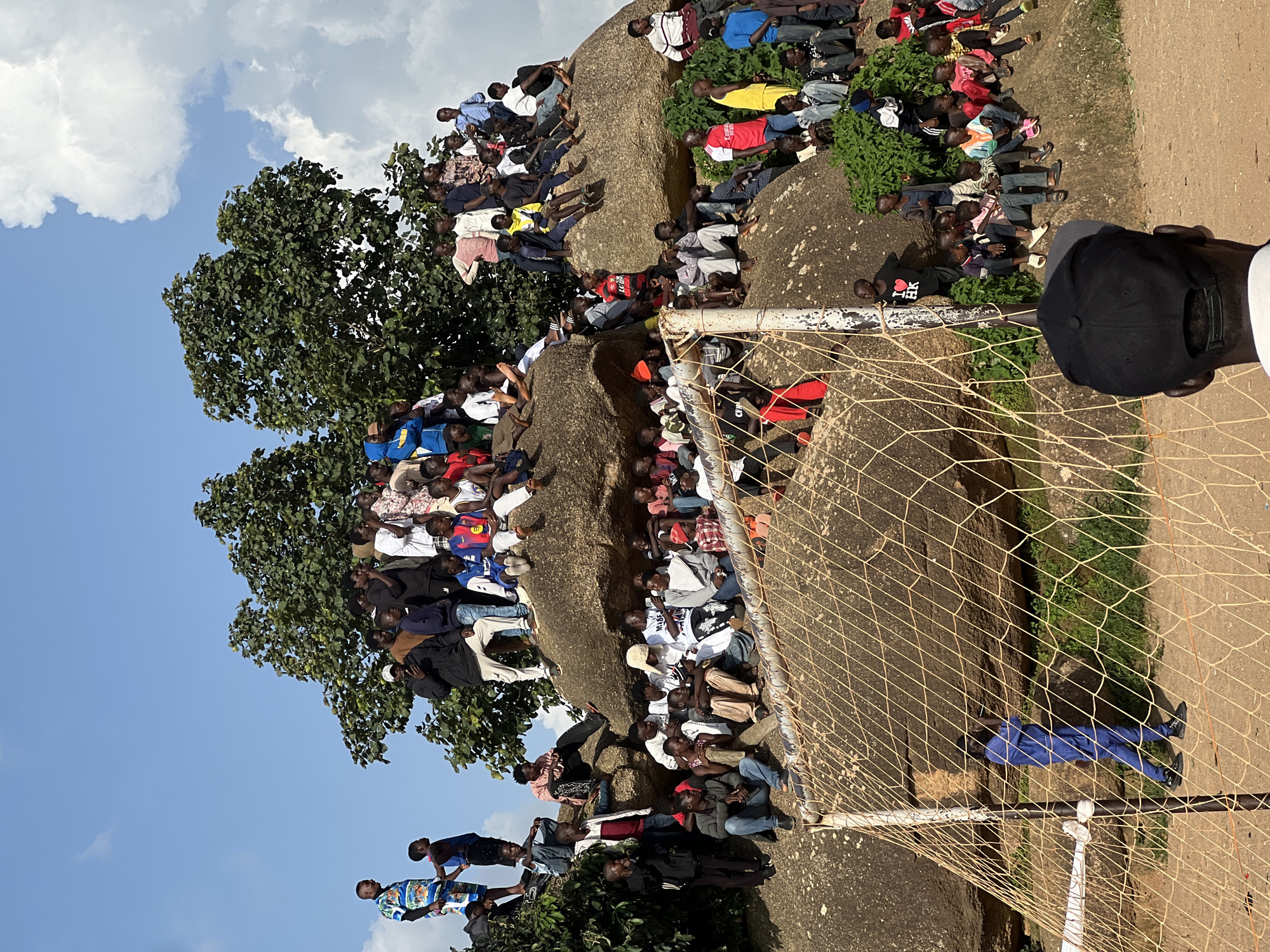 A large group of people are gathered on and around rocky outcrops, some sitting and others standing, with trees in the background under a cloudy sky. A soccer goal with a net is visible in the foreground. The scene appears to be outdoors, possibly a sporting event or social gathering, in Jos, Nigeria. The people are diverse in age and gender, with many dressed casually in t-shirts and shorts. The terrain is uneven, with large rocks dominating the landscape. The overall atmosphere seems to be relaxed and communal.