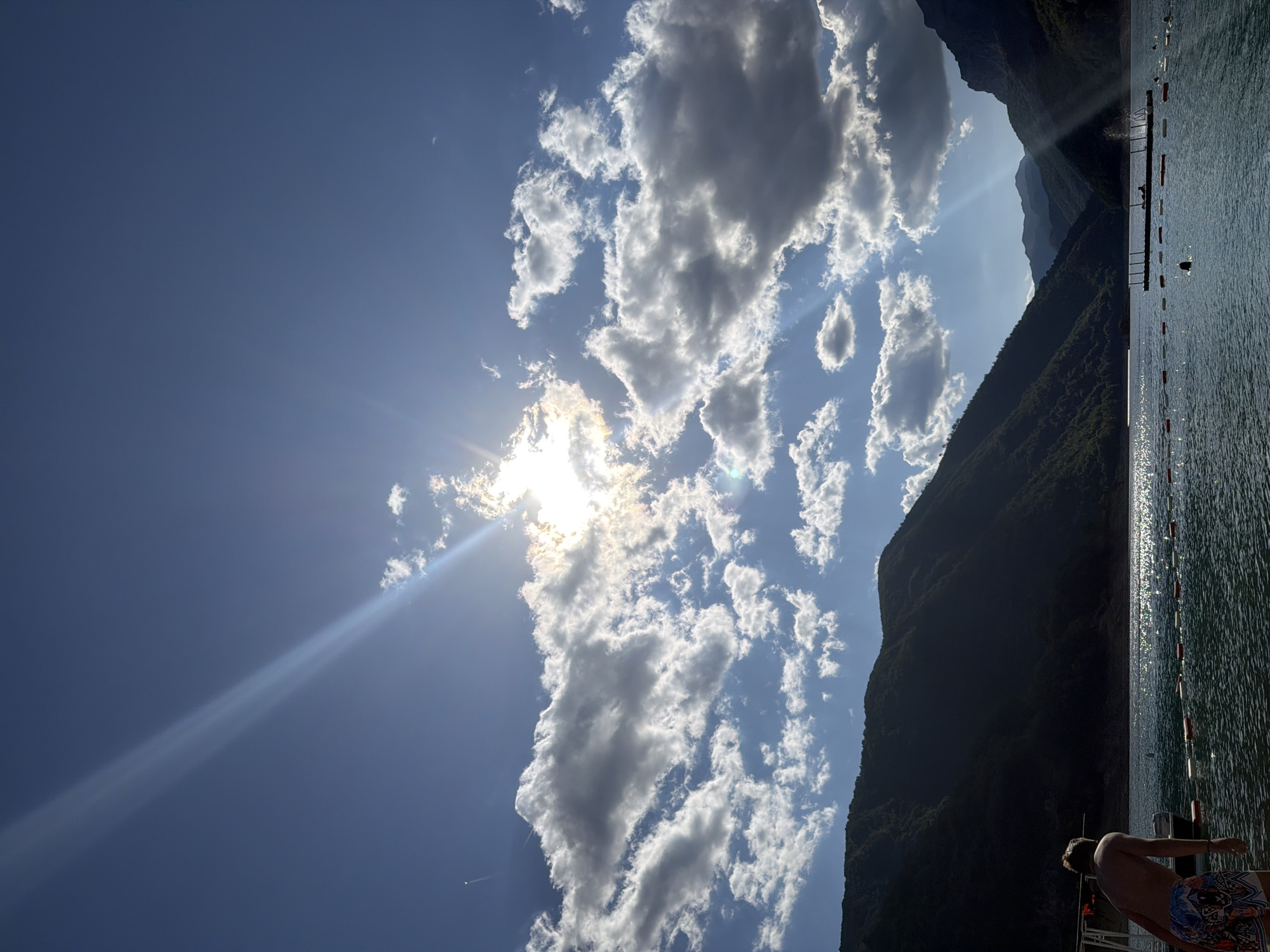 A wide shot captures a sun-drenched, mountainous landscape bordering a body of water, likely Piva Lake near Plužine, Montenegro. The sky is a vibrant blue, partially obscured by dramatic, white and grey cumulus clouds. The sun, a brilliant source of light, beams down, casting long rays that streak across the sky and glint off the water's surface. Majestic, dark green, tree-covered mountains flank the scene, their slopes descending towards the water. In the foreground, a young male, viewed from behind and wearing colorful patterned swim trunks, stands on a dock or shore, looking out at the water. The water itself is a deep blue, with a gentle ripple. Several buoys with red and white markings are visible, suggesting a designated swimming or recreational area. A small boat is moored to the left, and in the distance, a floating platform with some structures and people can be seen. The overall impression is one of a serene, outdoor recreational setting bathed in the warmth of a sunny day.