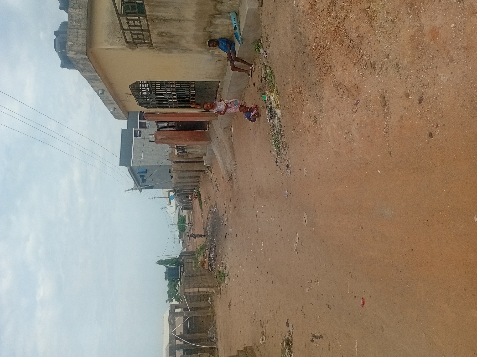 A street scene in Unknown, Unknown, shows a dirt road lined with buildings. Two children stand near a building, one holding a stick and the other sitting. Another child stands near a building further down the street. The sky is overcast.