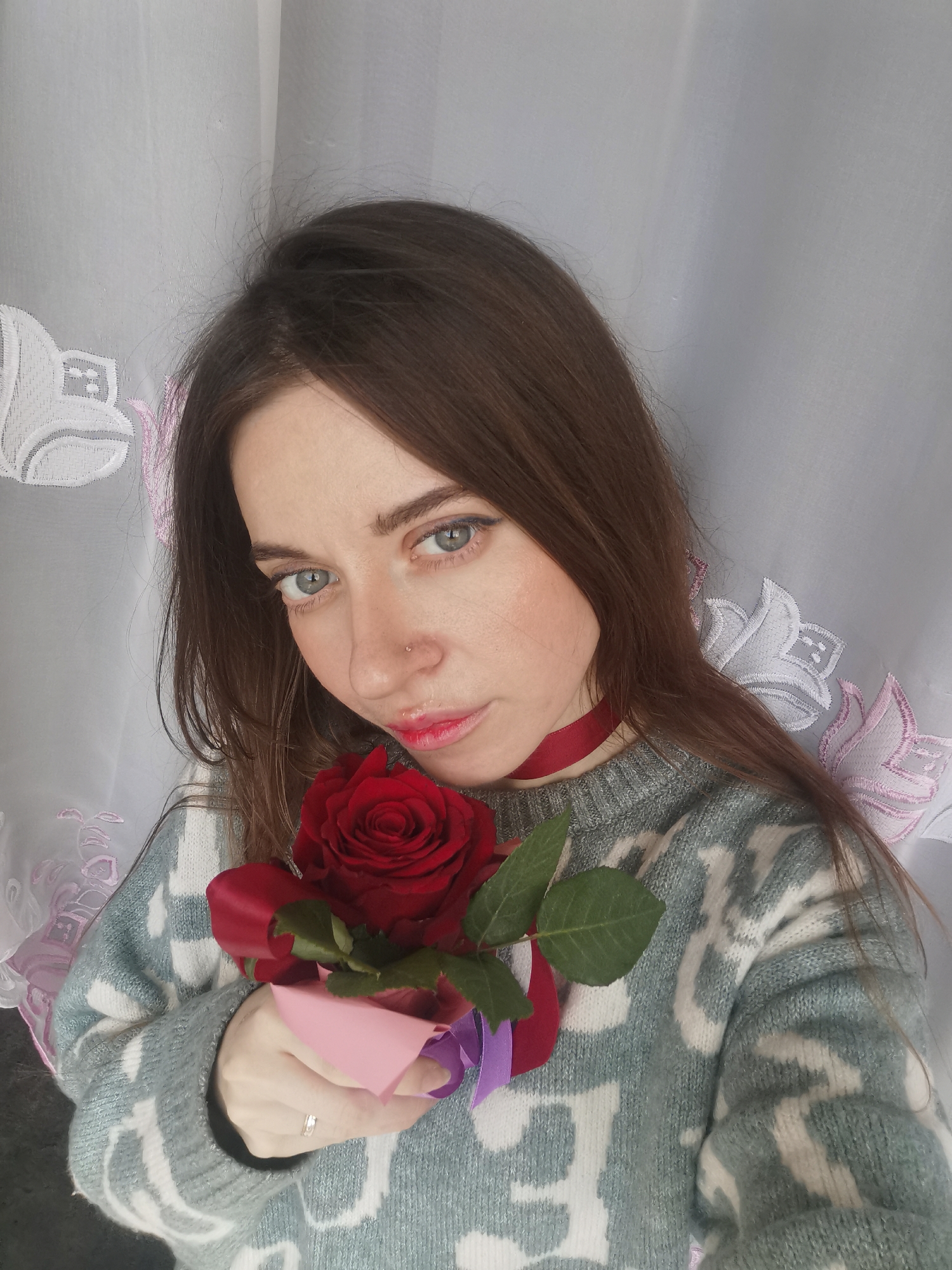 A head-and-shoulders portrait features a young woman with long, straight brown hair, blue eyes, and fair skin, looking directly at the camera. She has a small silver nose stud and is wearing red lipstick. A thin red ribbon is tied around her neck like a choker.
She is dressed in a light blue and white patterned knitted sweater. In her hands, positioned in front of her chest, she holds a single vibrant red rose, adorned with a wider red ribbon and partially wrapped in pink and purple paper. A silver ring is visible on her left ring finger.
The background consists of a sheer white curtain with embroidered floral patterns in white and soft pink. The lighting appears soft and natural, suggesting an indoor setting, possibly near a window. The woman's expression is neutral and direct. No text is visible in the image.