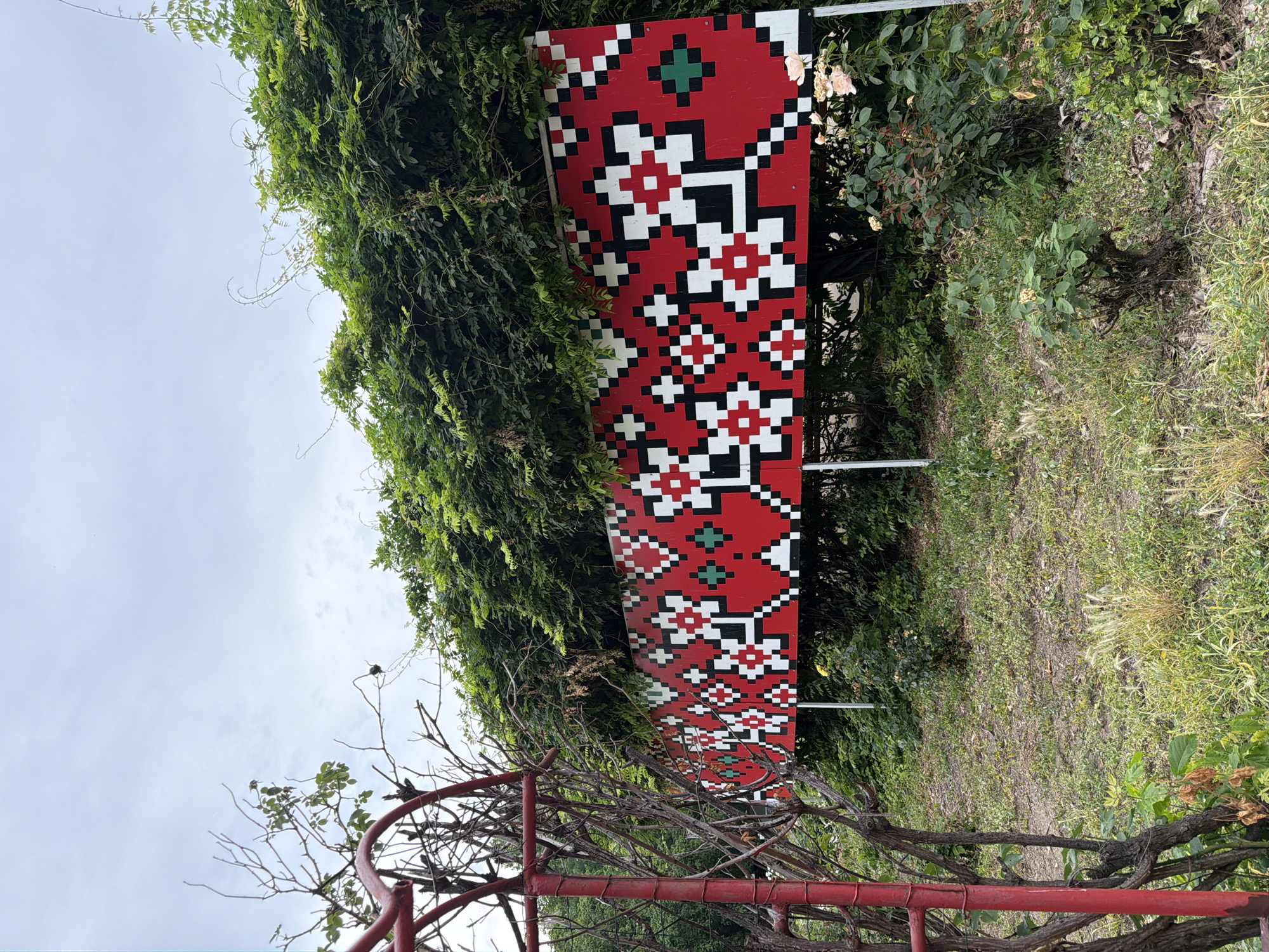 The image shows a rectangular banner with a red background and a pixelated geometric pattern in white, black, and green. The pattern resembles traditional folk embroidery. The banner is mounted on metal poles and is partially obscured by lush green foliage, suggesting it is outdoors. To the left, a red metal structure, possibly a trellis or part of a garden arch, is visible, along with dry branches and green leaves. The ground in front of the banner is covered with grass, dry leaves, and some small bushes with pink flowers. The sky above is overcast. The banner is located in the city of Pazardzhik, Bulgaria.