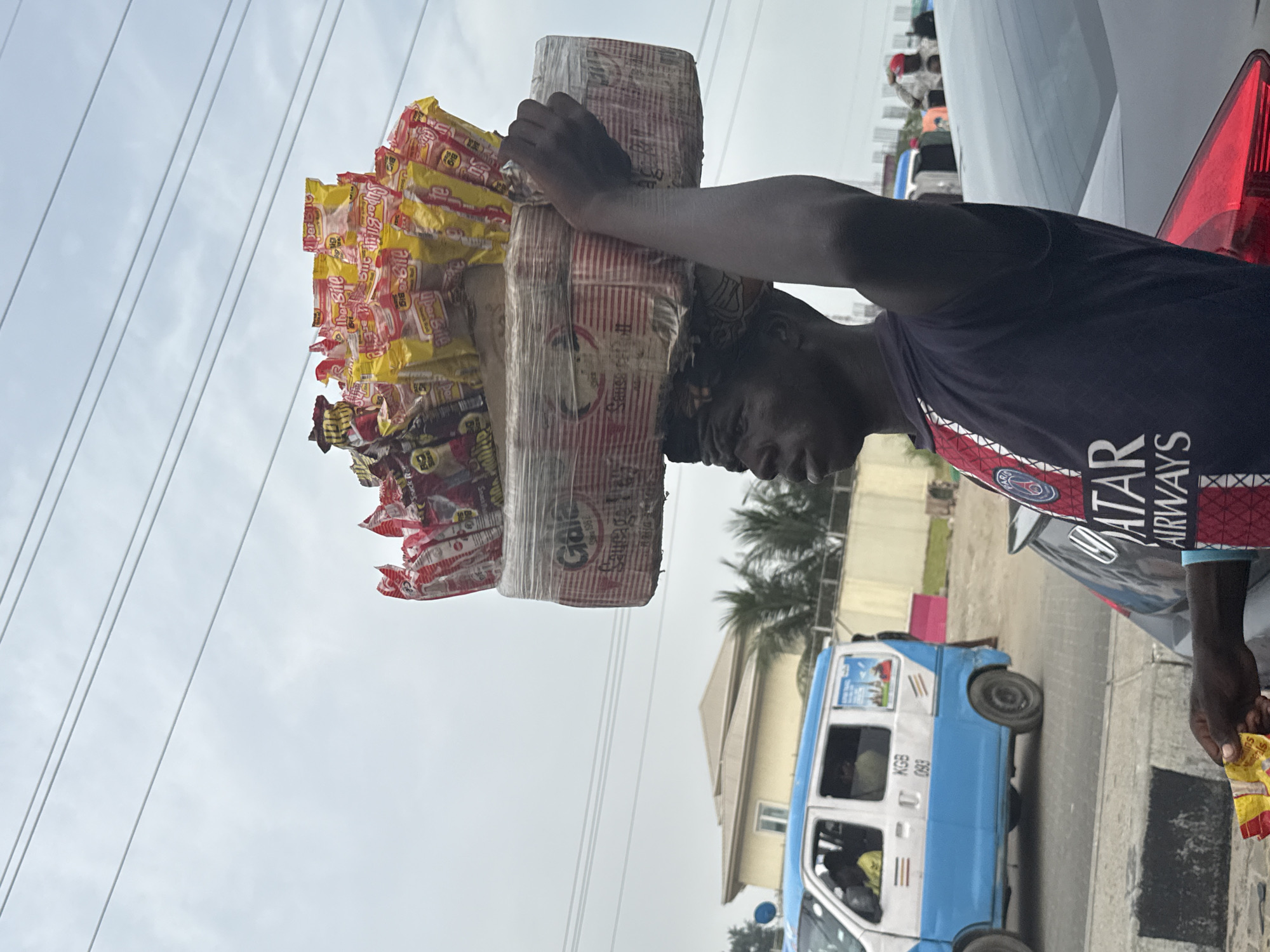The image shows a man in Alaguntan, Nigeria, carrying a large stack of packaged goods on his head. He is wearing a dark blue "QATAR AIRWAYS" soccer jersey and stands beside a car. The packages on his head appear to be snacks, with "Super Bite" visible on some. In the background, a light blue and white minivan with license plate "KGB 093" is parked. Power lines stretch across the sky. The area is likely a street with buildings and palm trees visible.