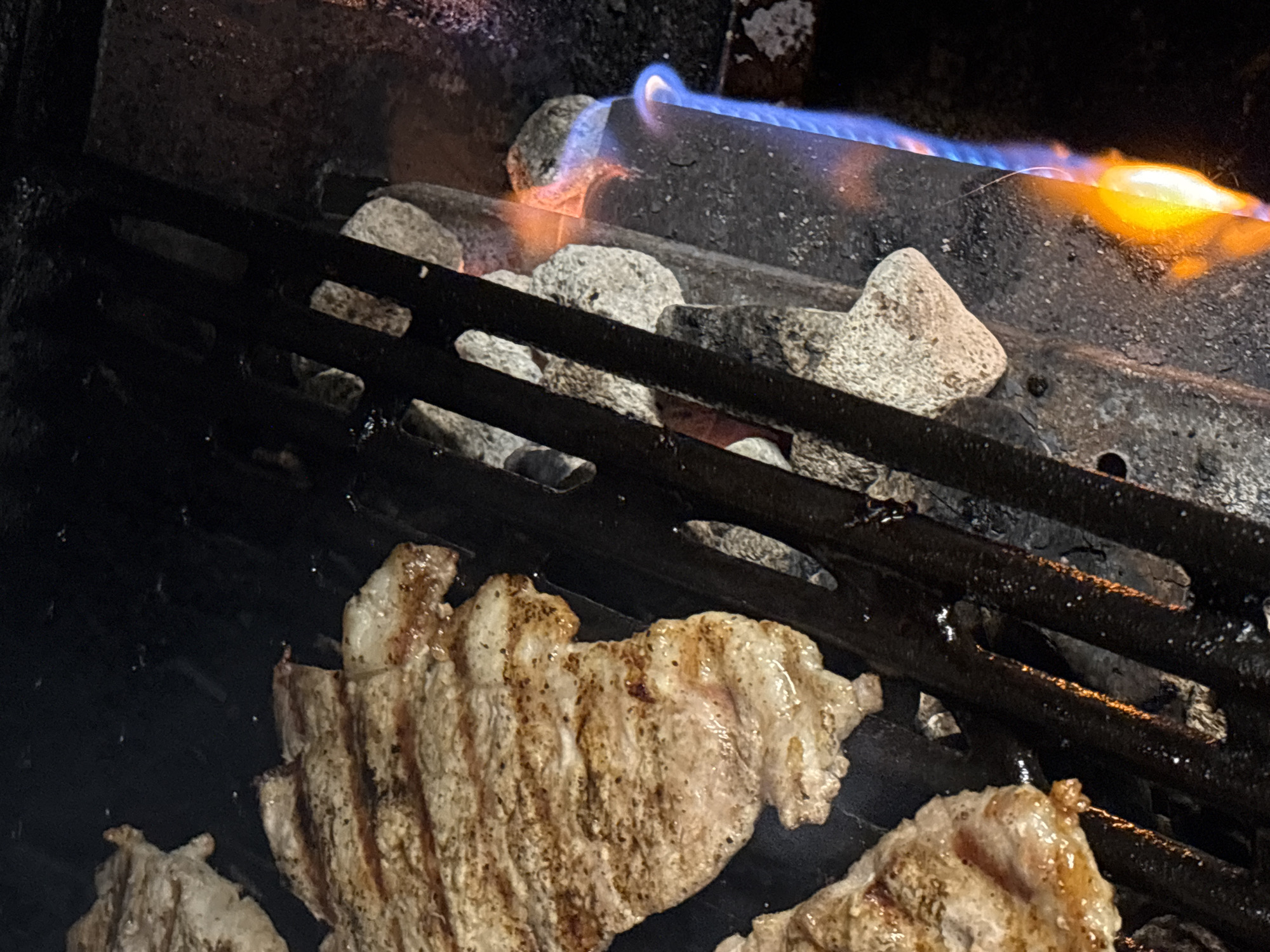 The image depicts food being grilled. Meat, possibly pork, is visible on a black metal grill. Above the grill, there are light-colored stones and a blue and orange flame, likely from a gas burner. The image lacks identifying details about a city or individuals; therefore, mentioning Lehchevo, Bulgaria, or details about people is not possible.