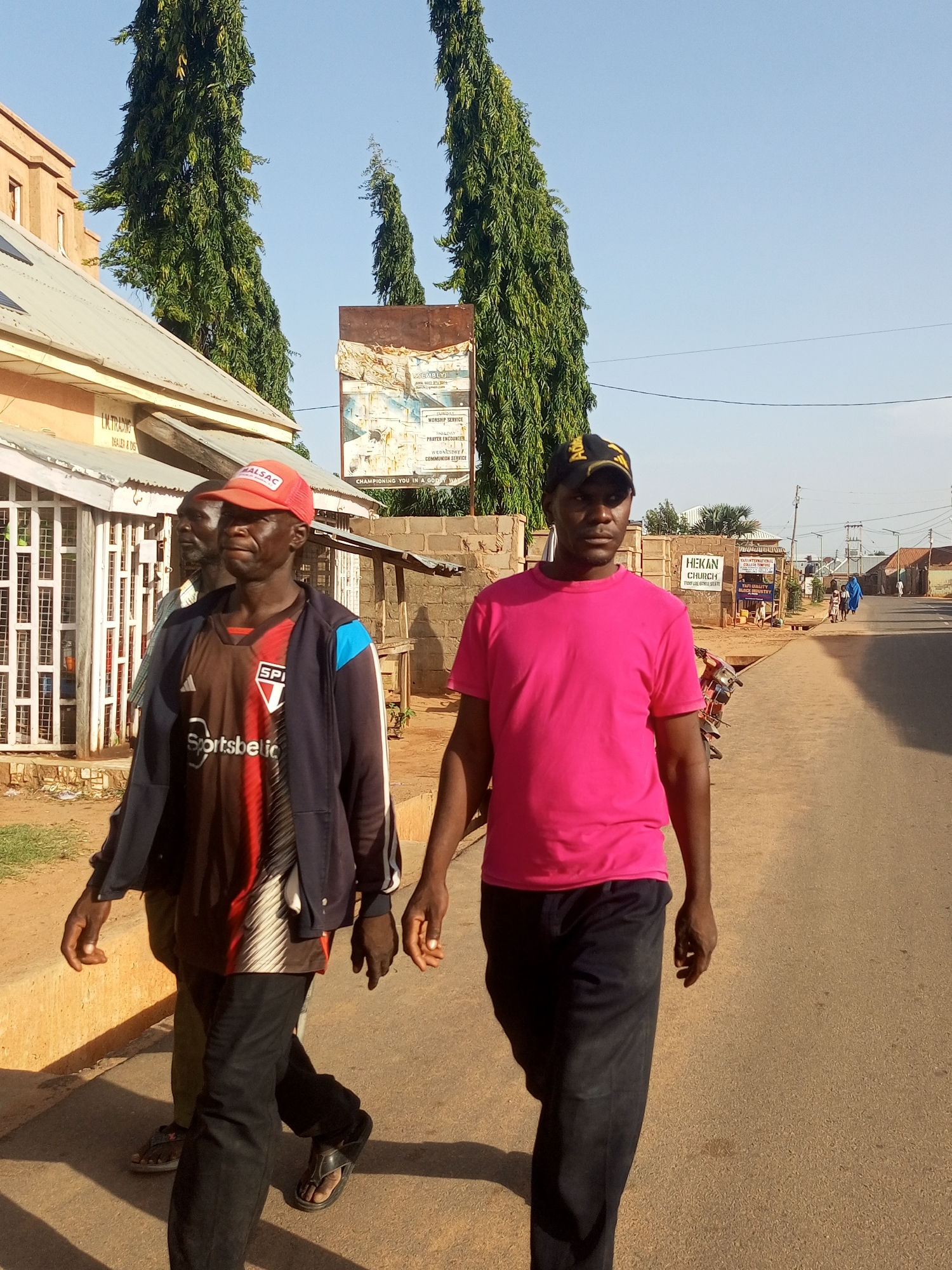 Two men are walking down a paved road in Gombe, Nigeria. The man in the foreground on the right is wearing a bright pink t-shirt and black pants, with a black baseball cap featuring a yellow logo. He is looking towards the camera with a neutral expression. To his left and slightly behind him is another man wearing a red baseball cap, a dark blue and red sports jersey, and black pants. He appears to be looking off to the side. A third person is partially visible behind them.
The scene is an outdoor, daytime setting under a clear blue sky. On the left side of the road, there are buildings with corrugated metal roofs and storefronts. One storefront has "I.M TRADING DEALER & DIG" visible. Further down the road, there are more buildings, including one with a sign that reads "HEKAN CHURCH". In the background, several tall, slender trees with dense green foliage stand out against the sky. A motorcycle is parked on the side of the road. The overall atmosphere appears to be casual and everyday. The presence of multiple shops and buildings suggests a commercial or residential area. The time of day appears to be late afternoon, given the angle of the shadows.