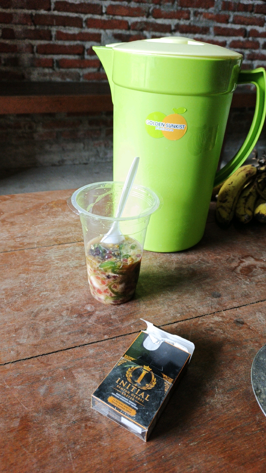 In Ngranti, Indonesia, on a weathered wooden table, rests a pack of "INITIAL" brand kretek cigarettes, open to reveal its foil lining. Next to it, a clear plastic cup holds a colorful beverage with visible chunks of fruit and possibly herbs, a white plastic spoon submerged within. Behind the cup stands a large, bright green pitcher, its lid on and a "GOLDEN SUNKIST plasticware" sticker adhered to its side. A bunch of ripe bananas lies to the right of the pitcher. The background features a brick wall and wooden shelving.